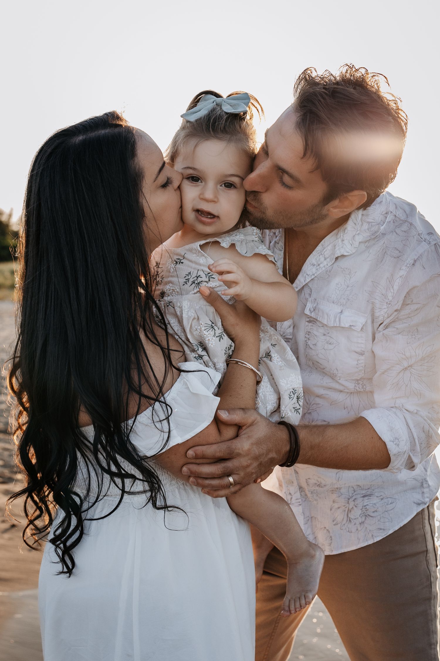 Parents kissing their baby girl on cheeks at golden hour beach.
