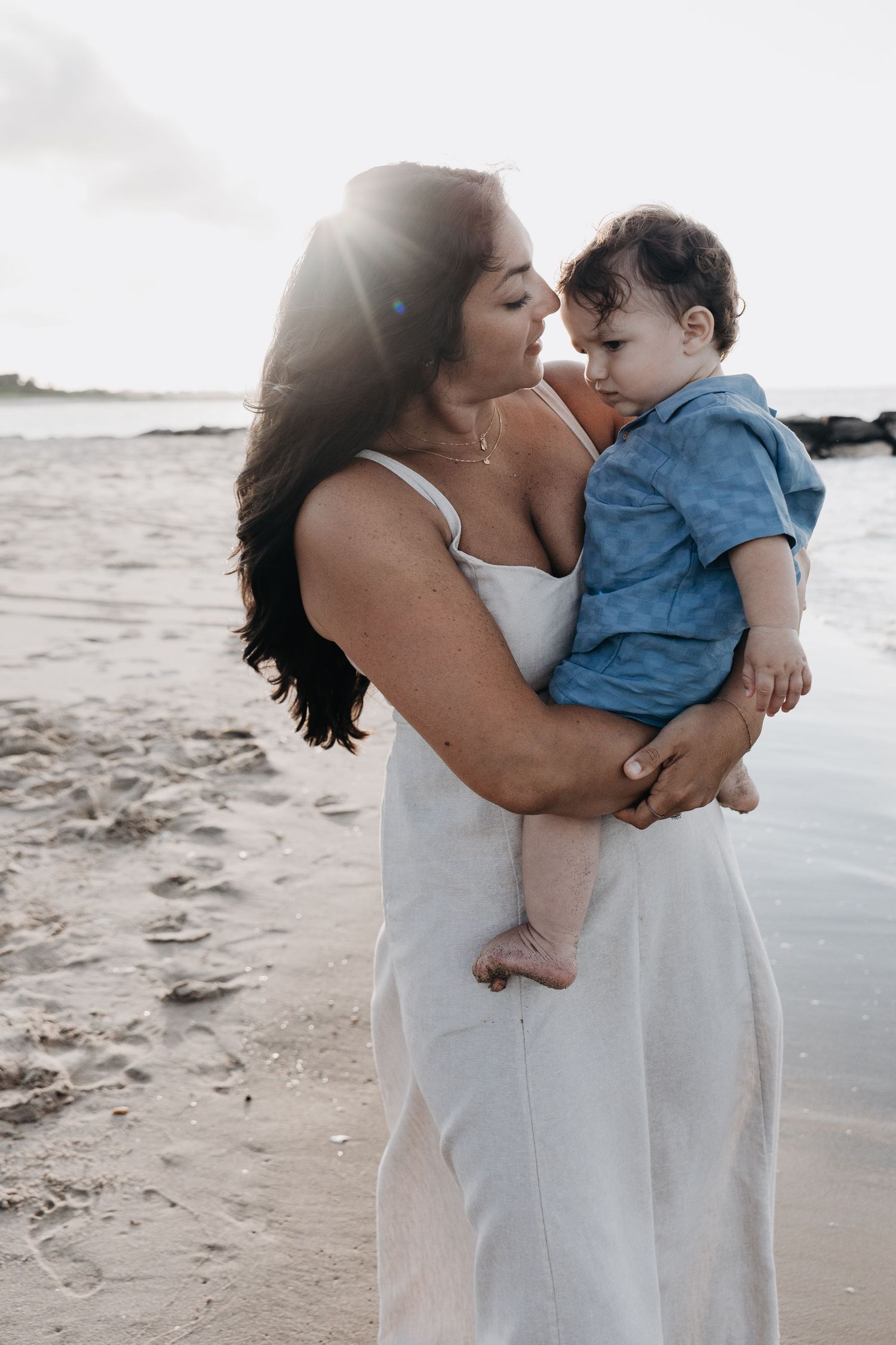 Mother in white dress holding baby boy on sunny beach at sunset.
