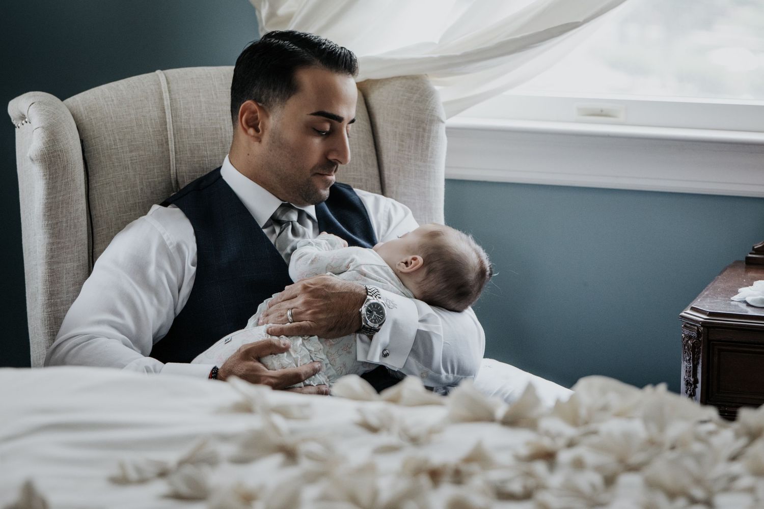 Formally dressed man sitting in chair tenderly holding sleeping baby in christening gown.