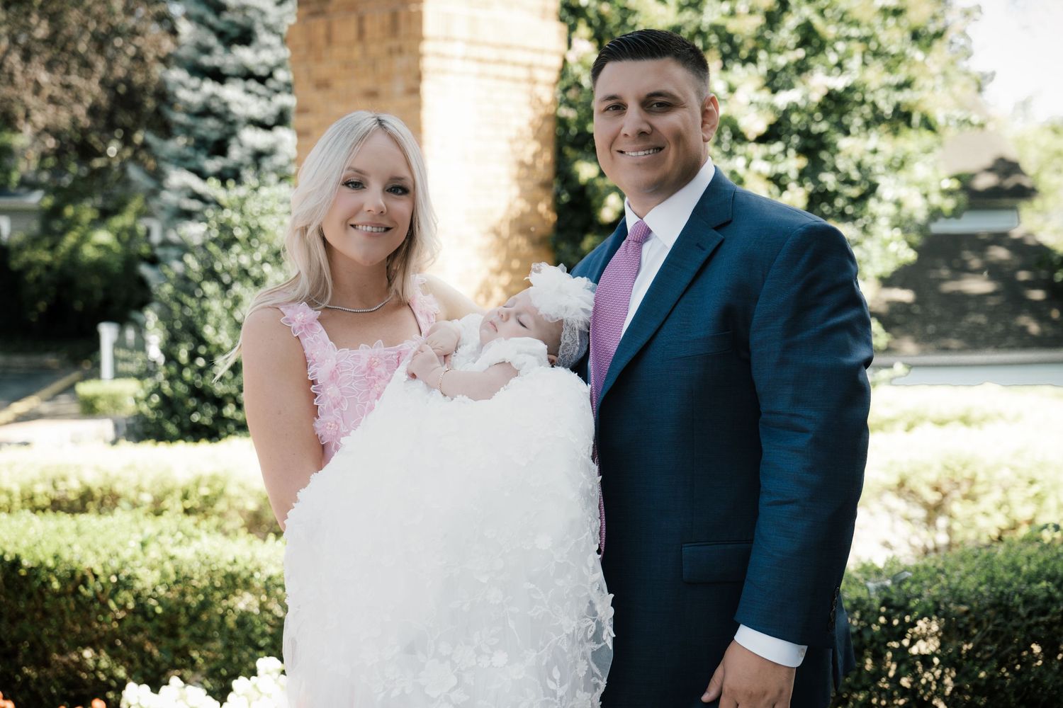 Smiling couple in formal attire holding baby in white christening gown outdoors.