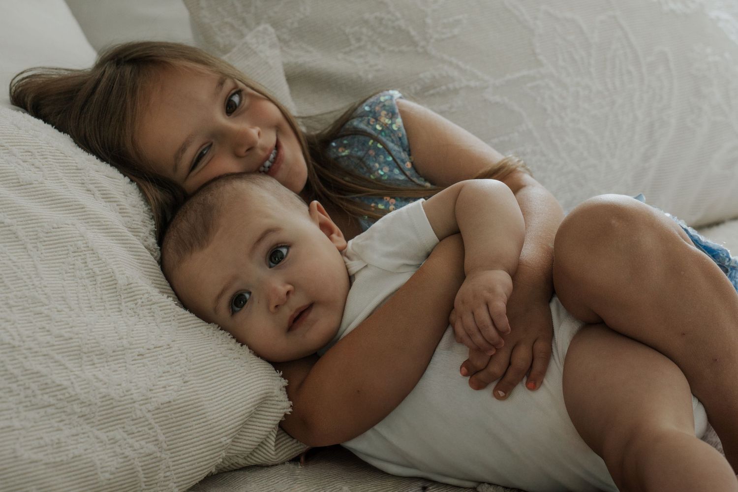 Older sister lovingly hugging baby sibling on white bedding.
