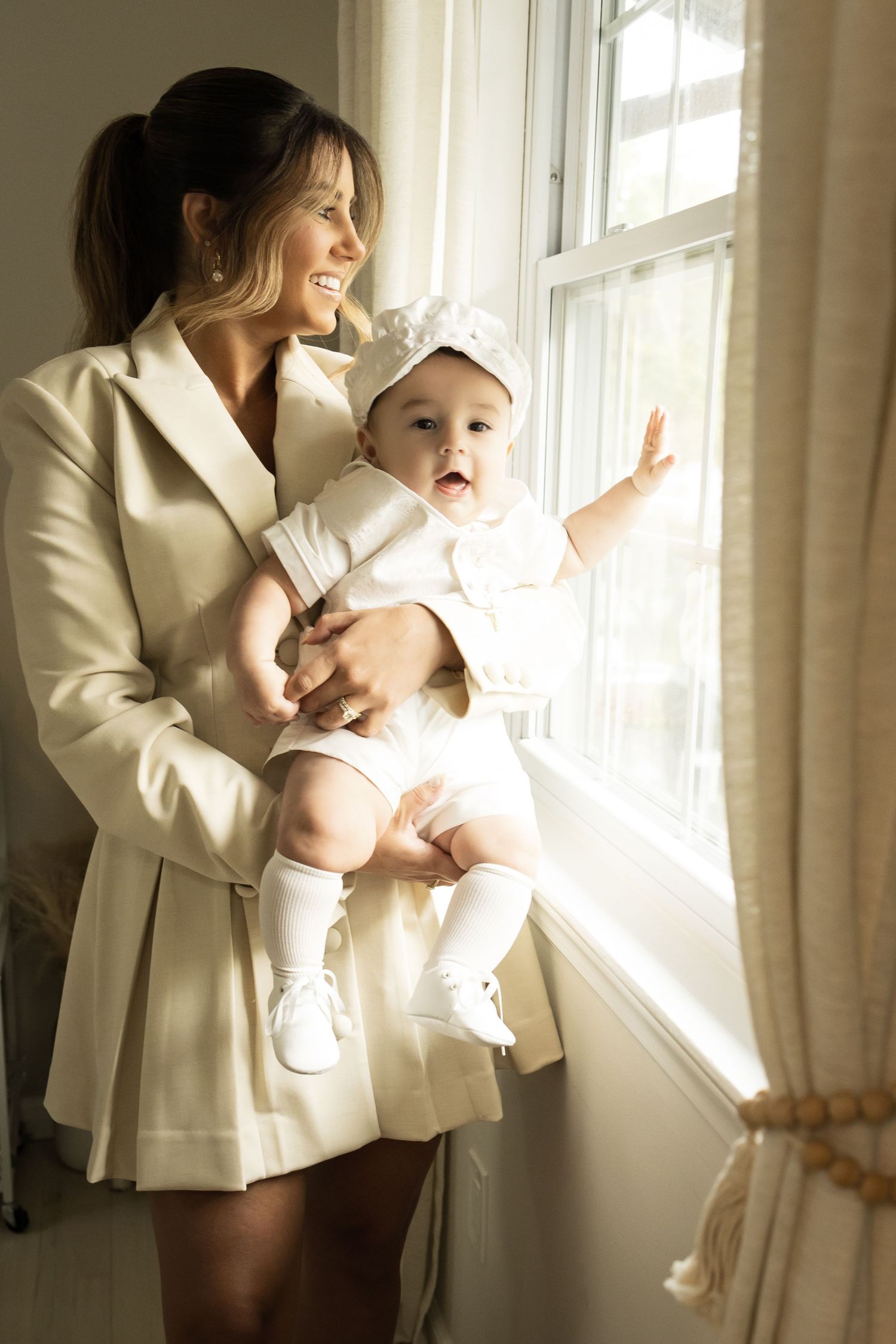 Smiling mother in cream dress holding baby in white christening outfit near sunlit window.