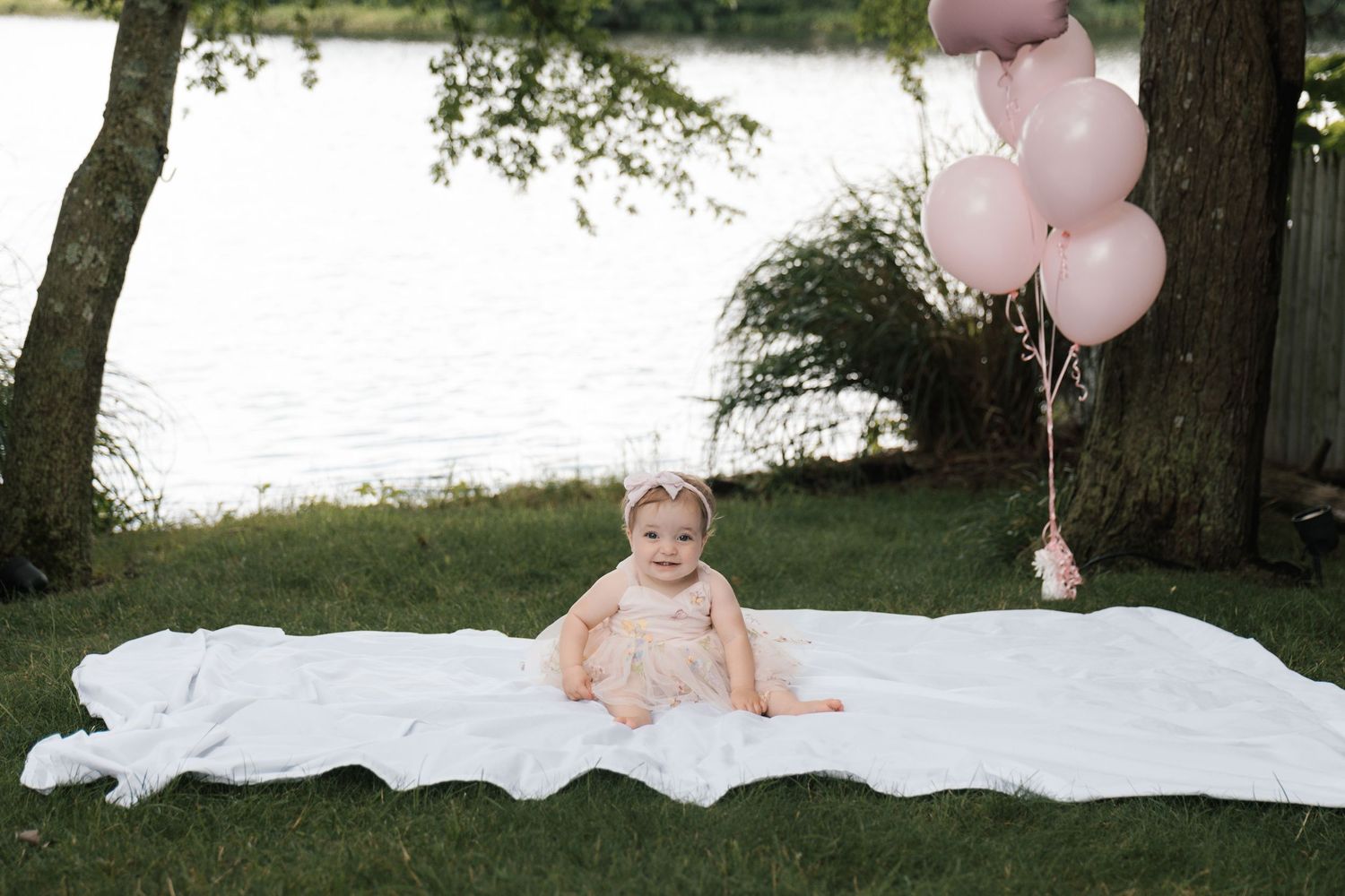 Baby girl in pink dress sits on white blanket near pink balloons by lake.