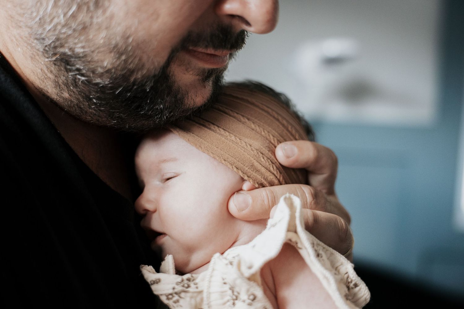 Father with beard tenderly holds sleeping newborn baby against his chest in soft natural light.