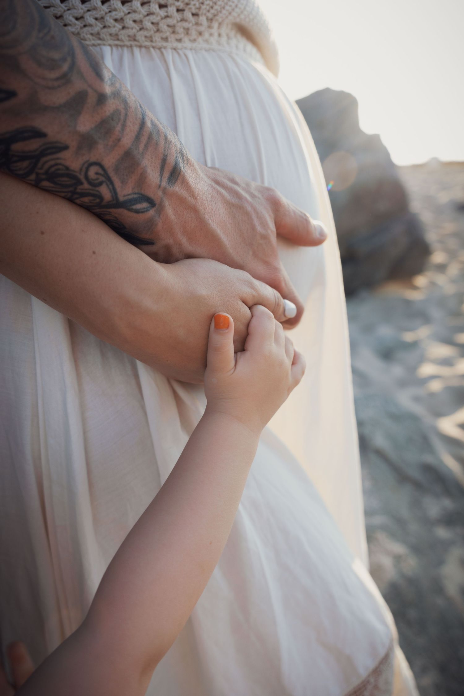 Child holding tattooed fathers hand near pregnant mothers belly at beach.