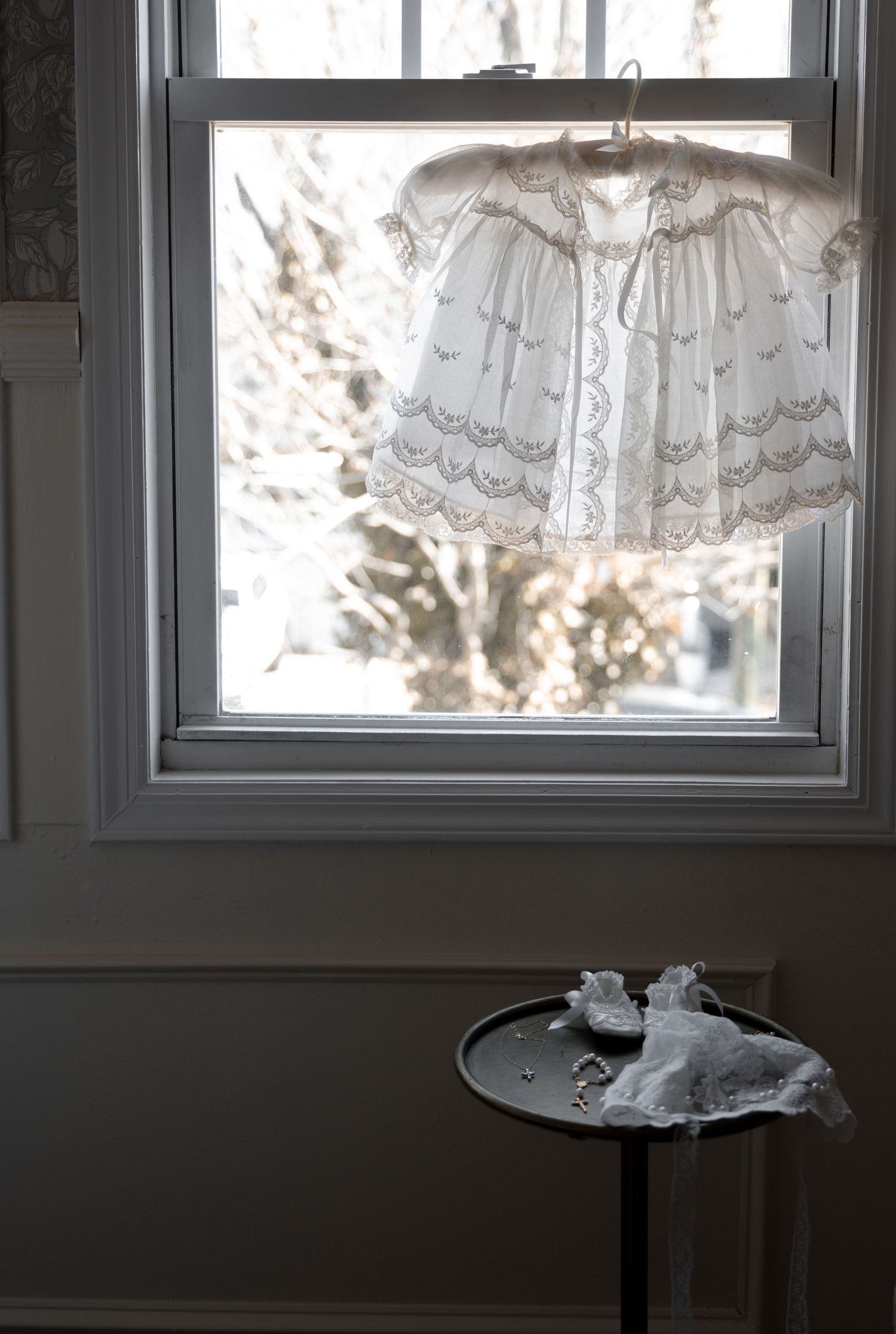 Delicate white lace christening gown hanging in a sunlit window above a small antique side table.