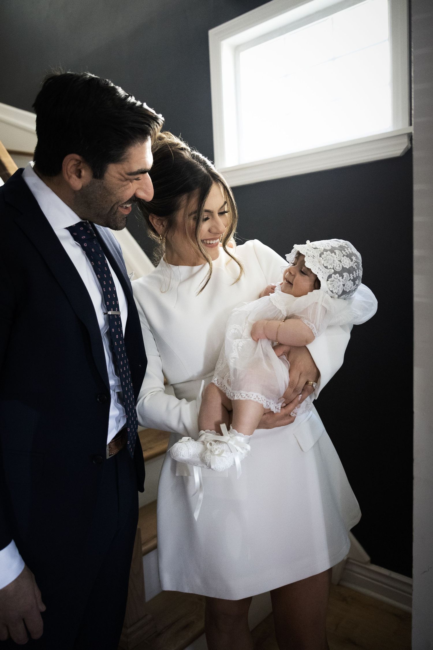 Smiling parents in formal wear lovingly admiring their baby dressed in white christening gown and bonnet.