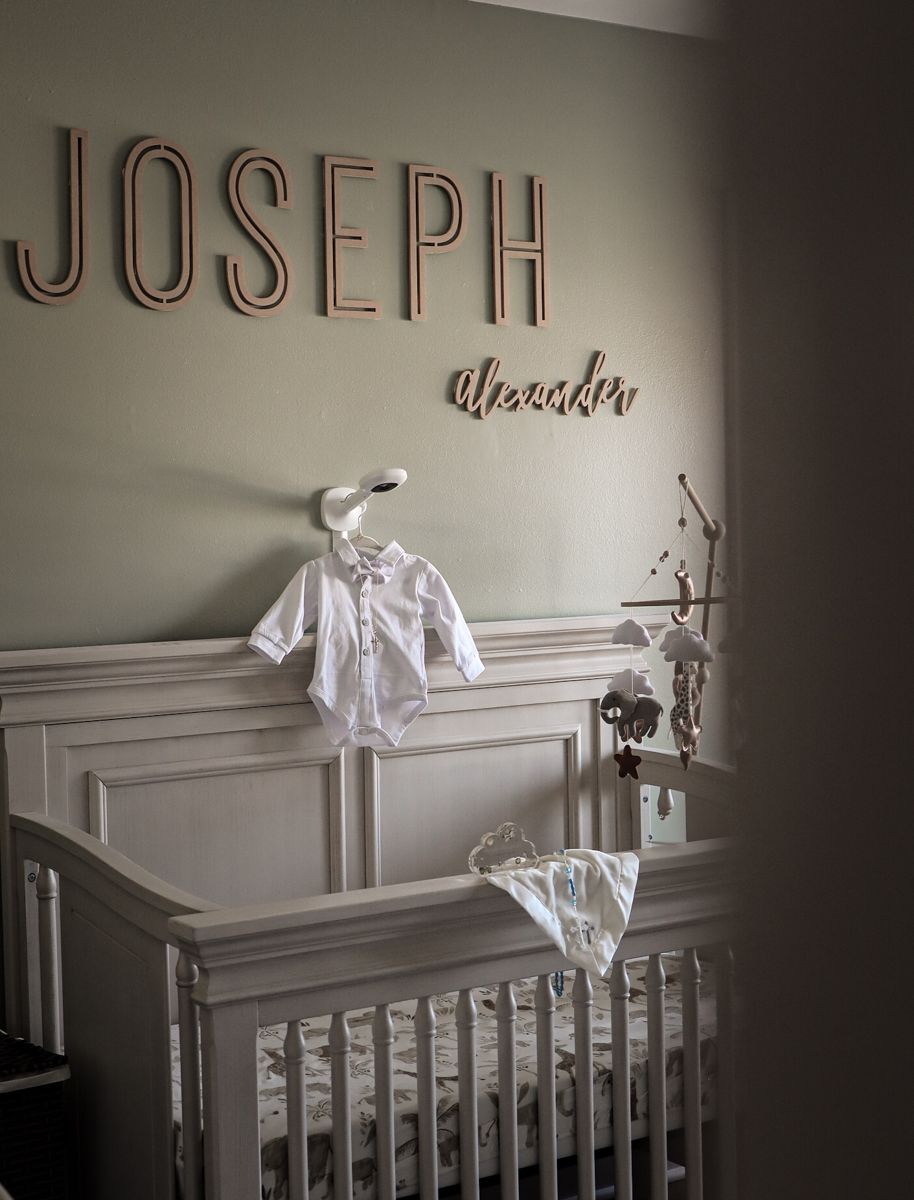 Elegant nursery with Joseph Alexander wooden name letters above a white crib with baby clothes displayed.