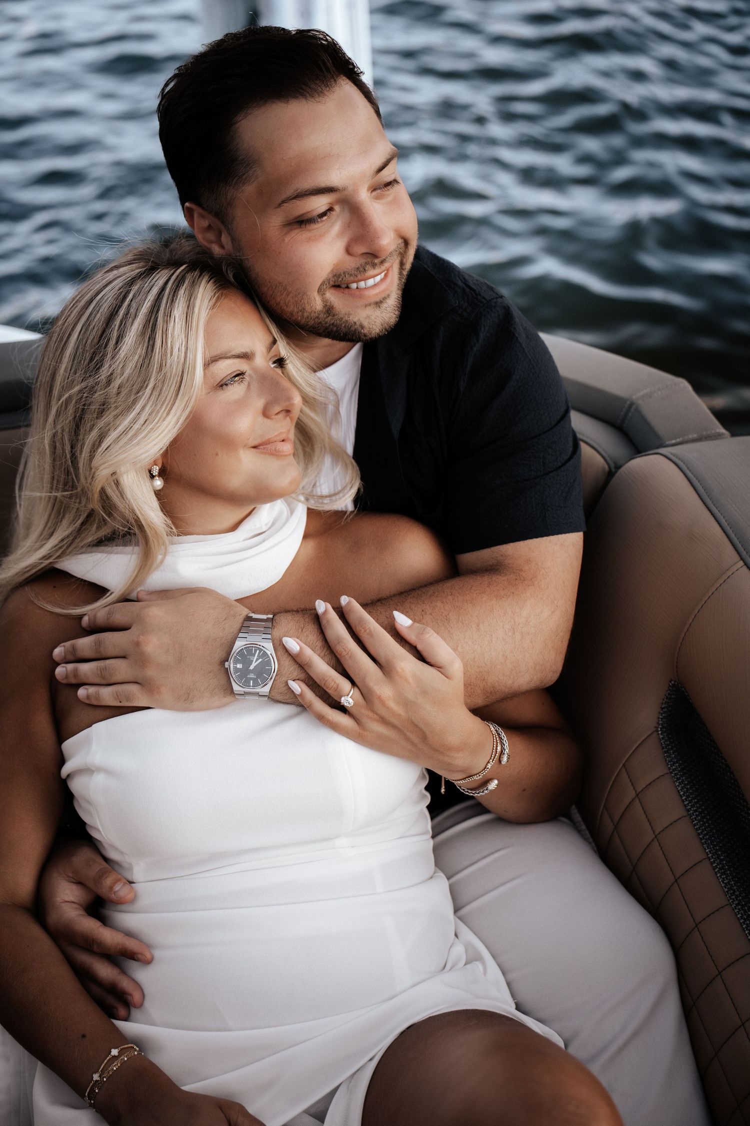Man hugging woman in white dress on a boat with water backdrop during engagement shoot.