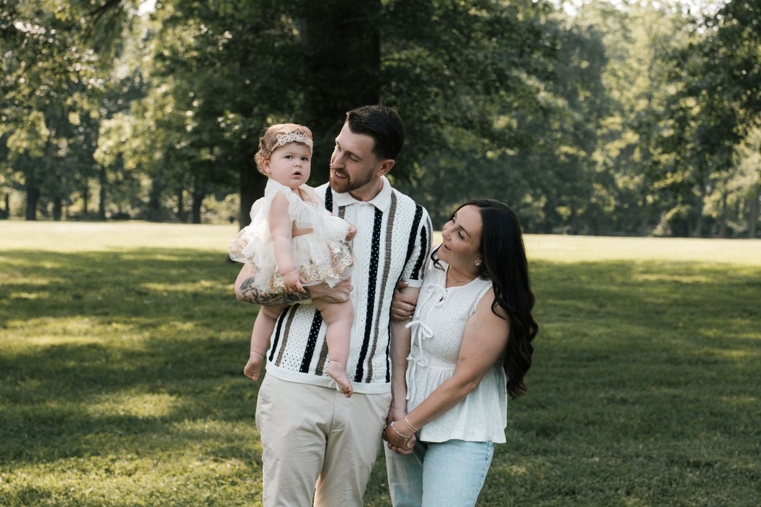A couple smiles in a sunny park while the man holds their young daughter in a pink dress.