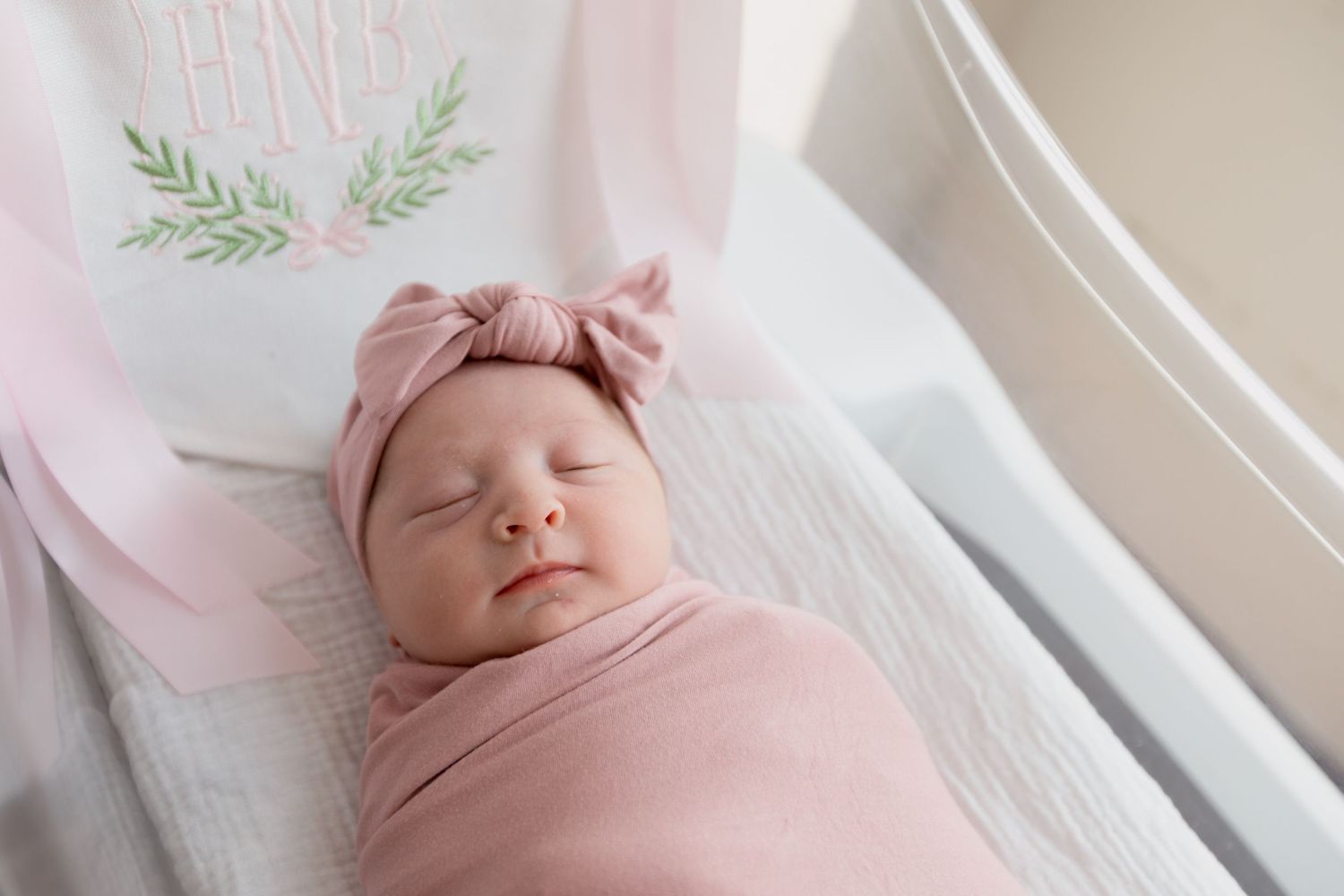 A sleeping newborn baby wrapped in soft pink swaddle blanket with bow headband rests peacefully on white textured bedding.