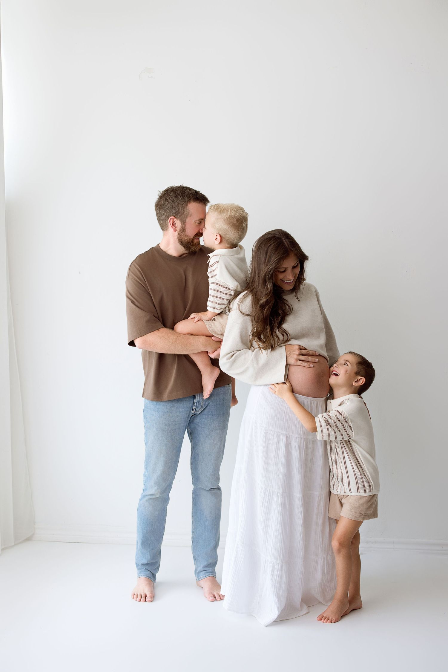 A family wearing neutral colors embraces each other lovingly in a bright white studio setting.