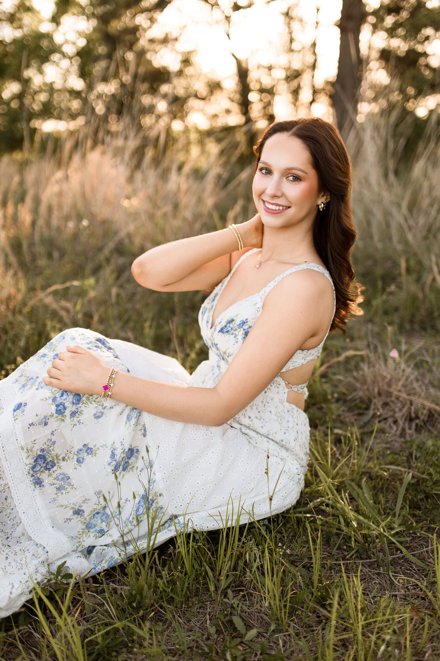 Young woman in white floral dress sitting in golden hour sunlight among tall grass, smiling gracefully.