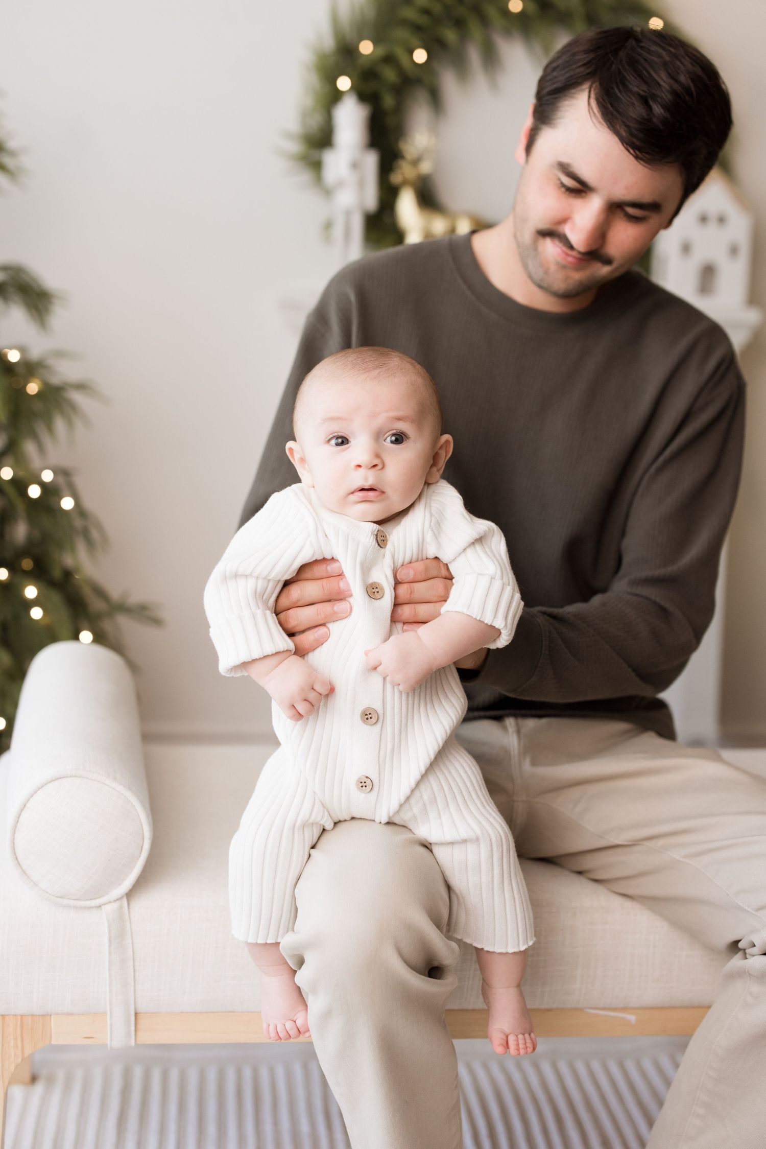 A series of four photos showing a baby in white knit clothing sitting with an adult in a cozy holiday-themed setting.