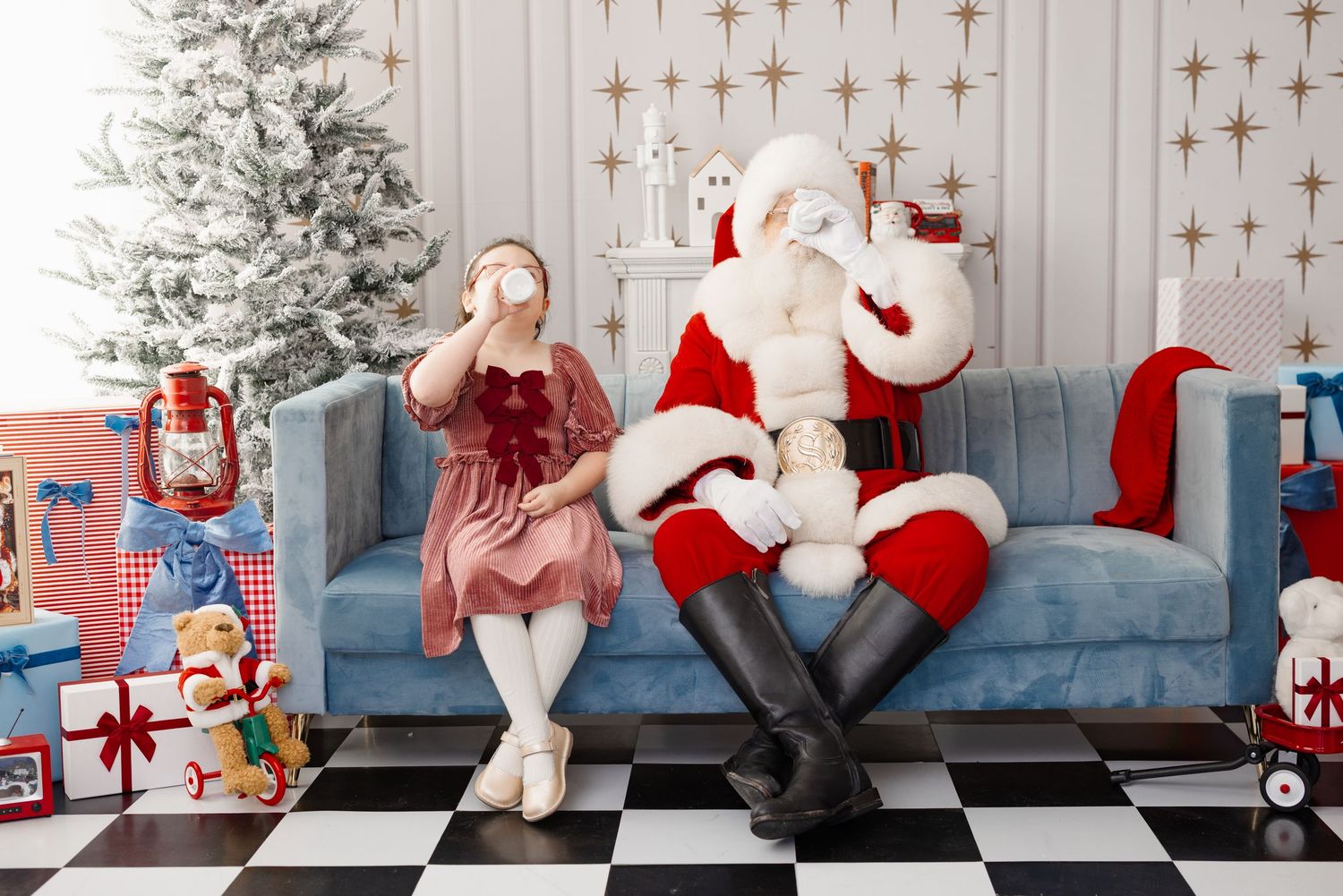 A festive holiday scene with Santa seated on a blue couch next to a decorated Christmas tree with presents and star decor.