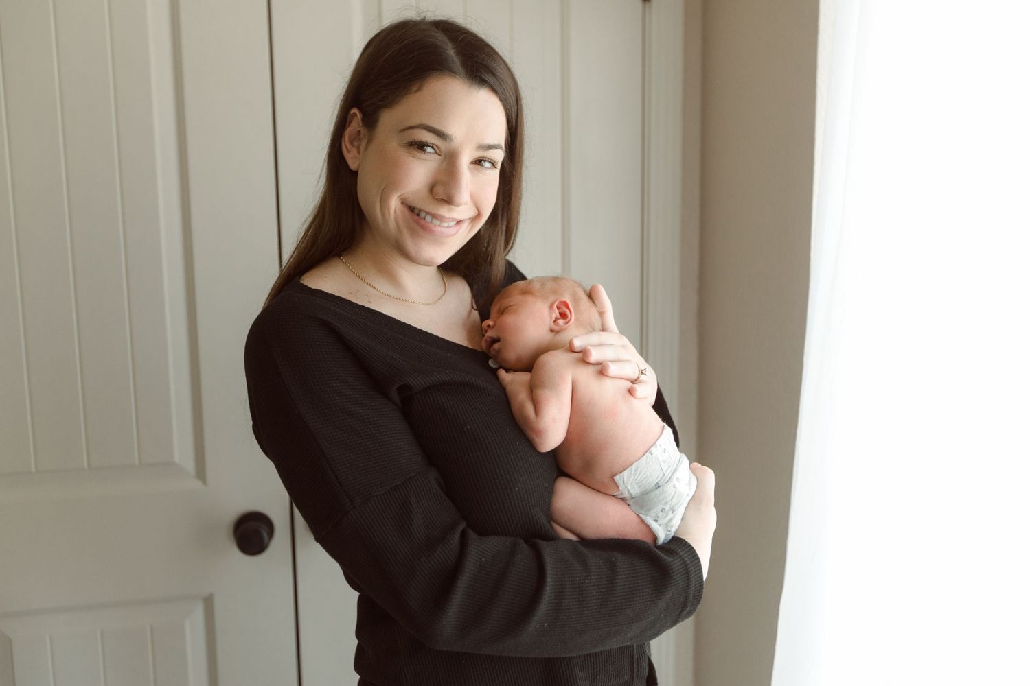 A parent in a black sweater holds a sleeping newborn baby against their shoulder in a softly lit room during Fresh 48 nursery session.