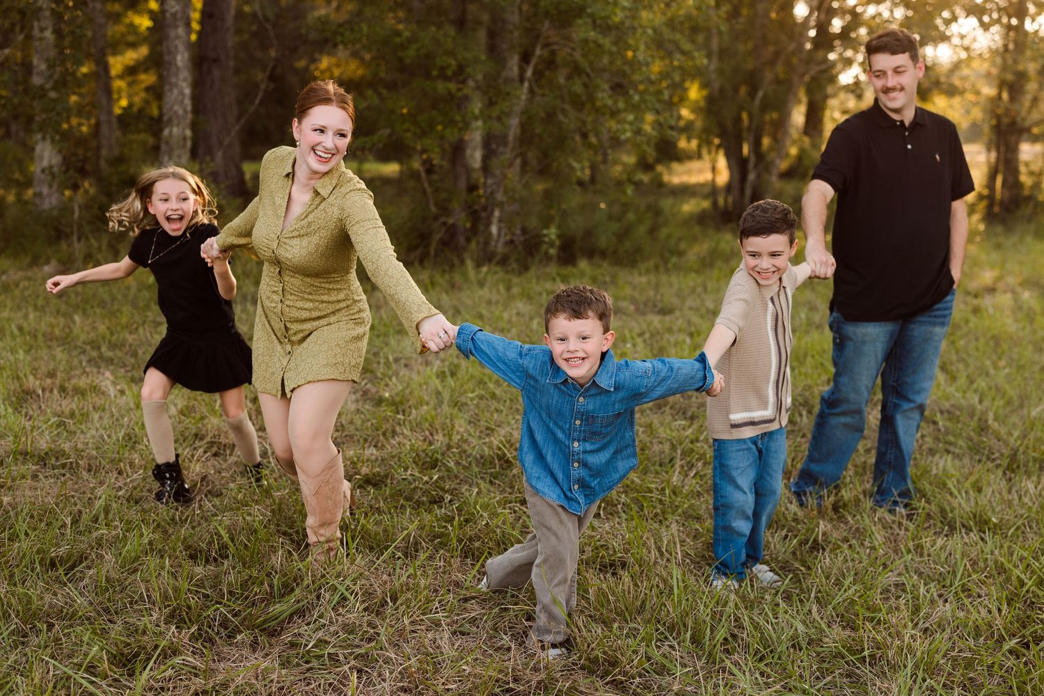 Smiling family of five holding hands and playing together in a sunny grassy field with tall trees in the background.