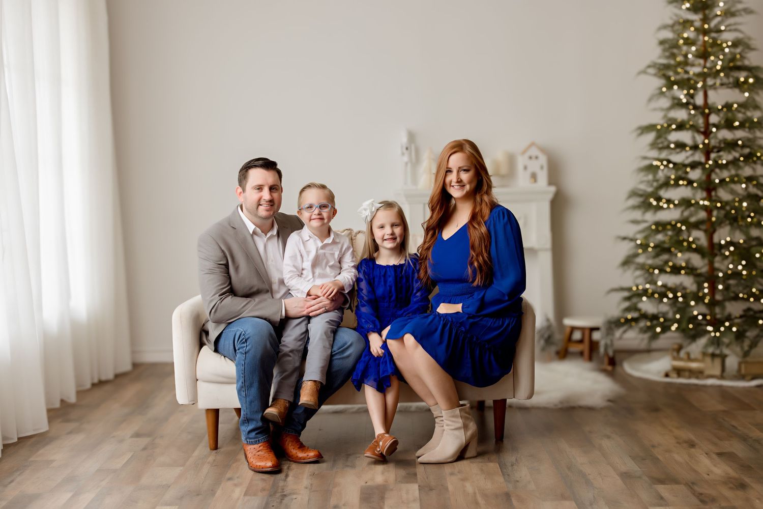 A family of four in coordinating blue and gray outfits pose together on a white couch near a Christmas tree.