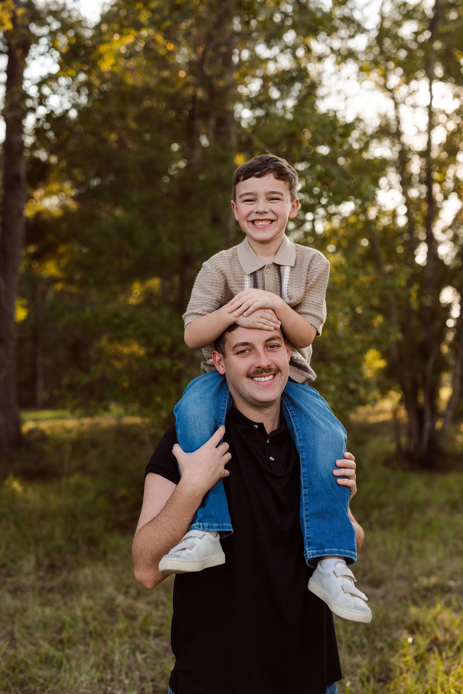 Smiling young boy sitting on the shoulders of a mustached man in a black polo, standing in a sunlit grassy park surrounded by trees.