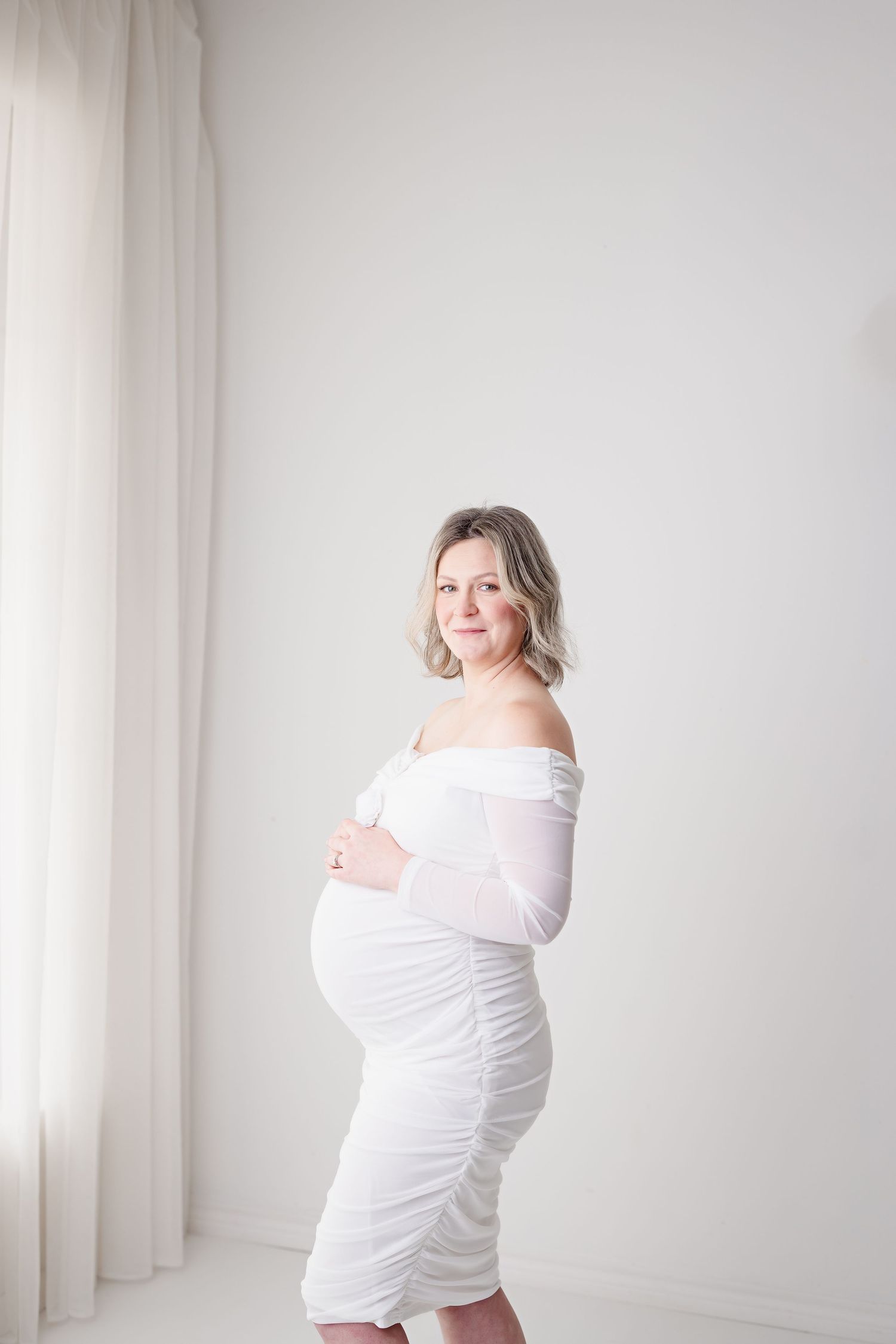 Ethereal maternity portrait in white ruched dress beside sheer curtains with Bree Elle Photography in Kountze, TX.