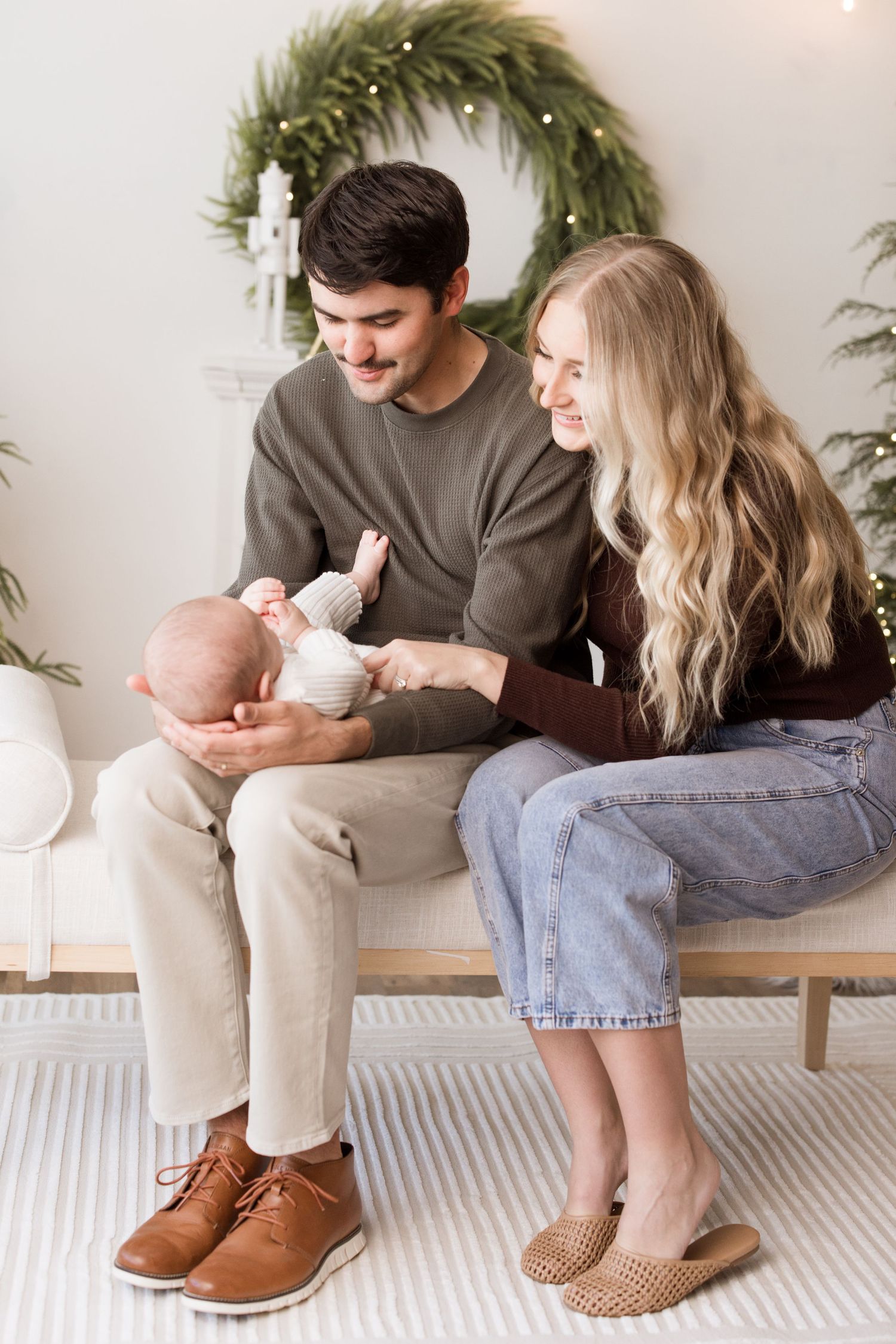 Young couple sitting on bench with baby under holiday wreath in cozy indoor setting.