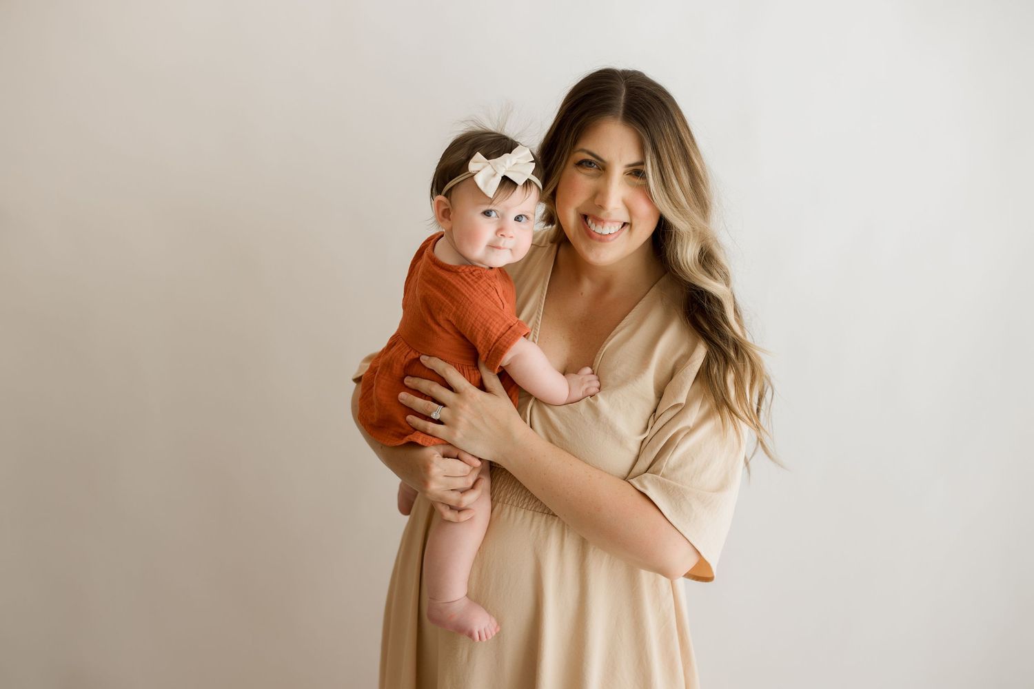 Person in beige dress holds small child wearing orange outfit against neutral background.