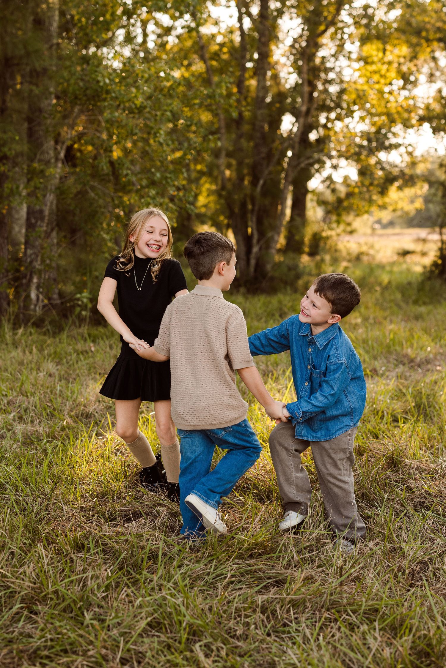 Three smiling children hold hands and play in a circle on a grassy field with sunlit trees in the background.