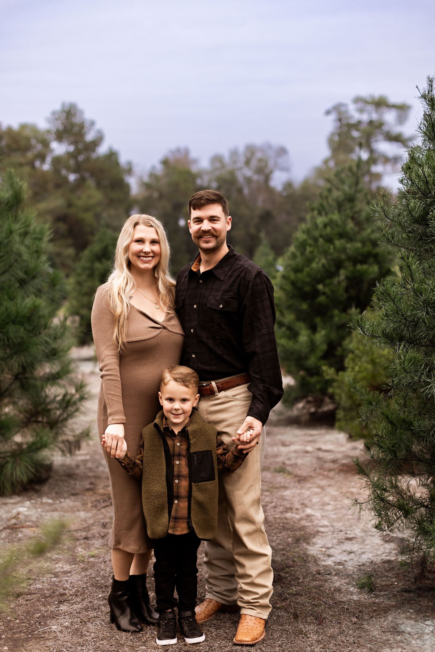 A family poses together on a path surrounded by evergreen trees at a Christmas tree farm.