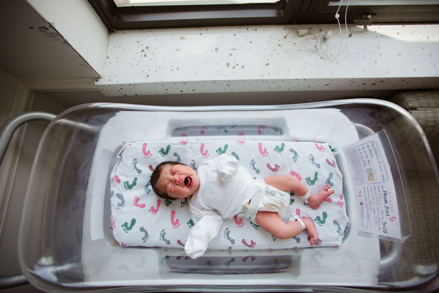 Newborn baby lying in a clear hospital bassinet on a patterned sheet.