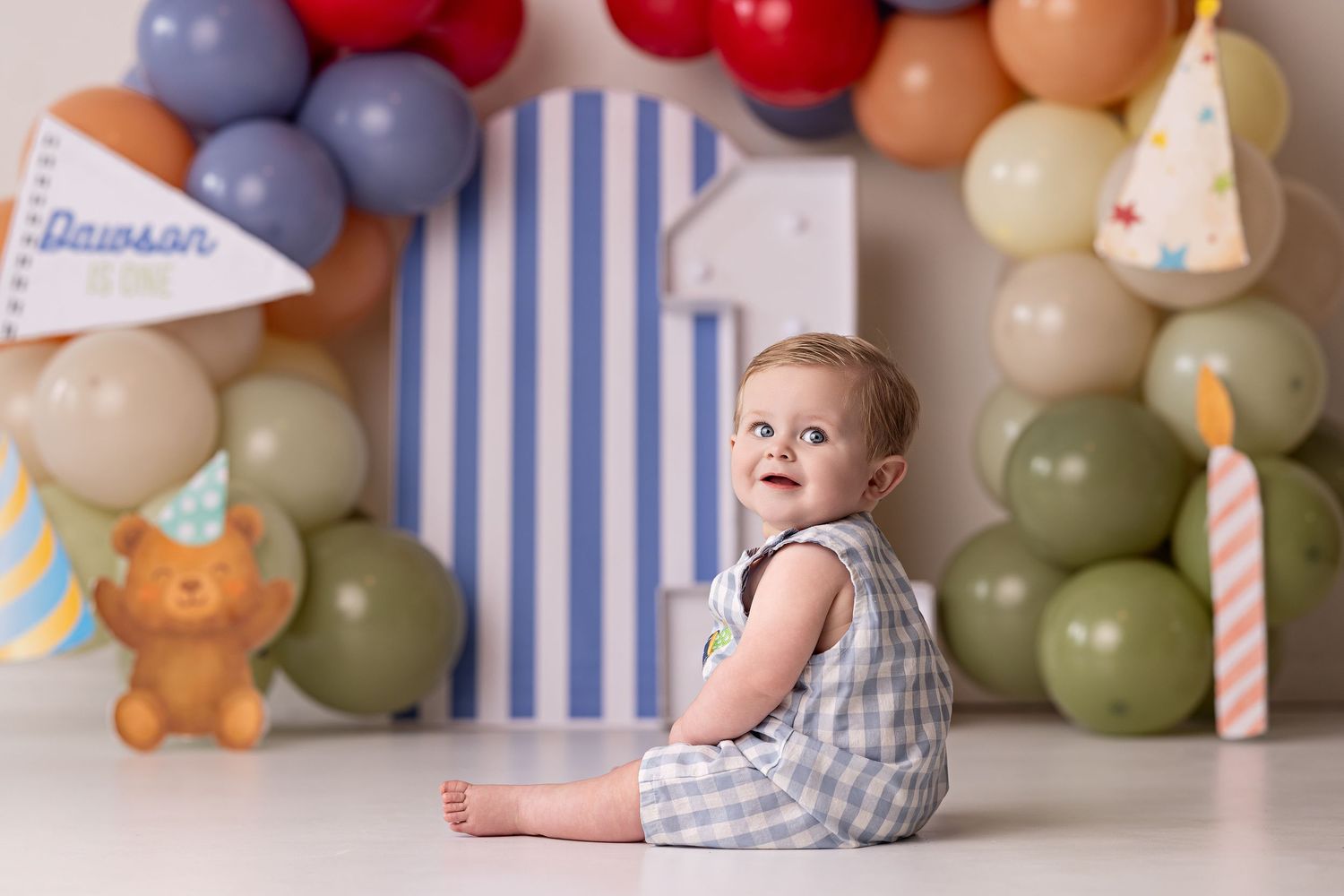 A baby in a grey outfit sits on a wooden floor with colorful balloon decorations and striped wallpaper in the background.
