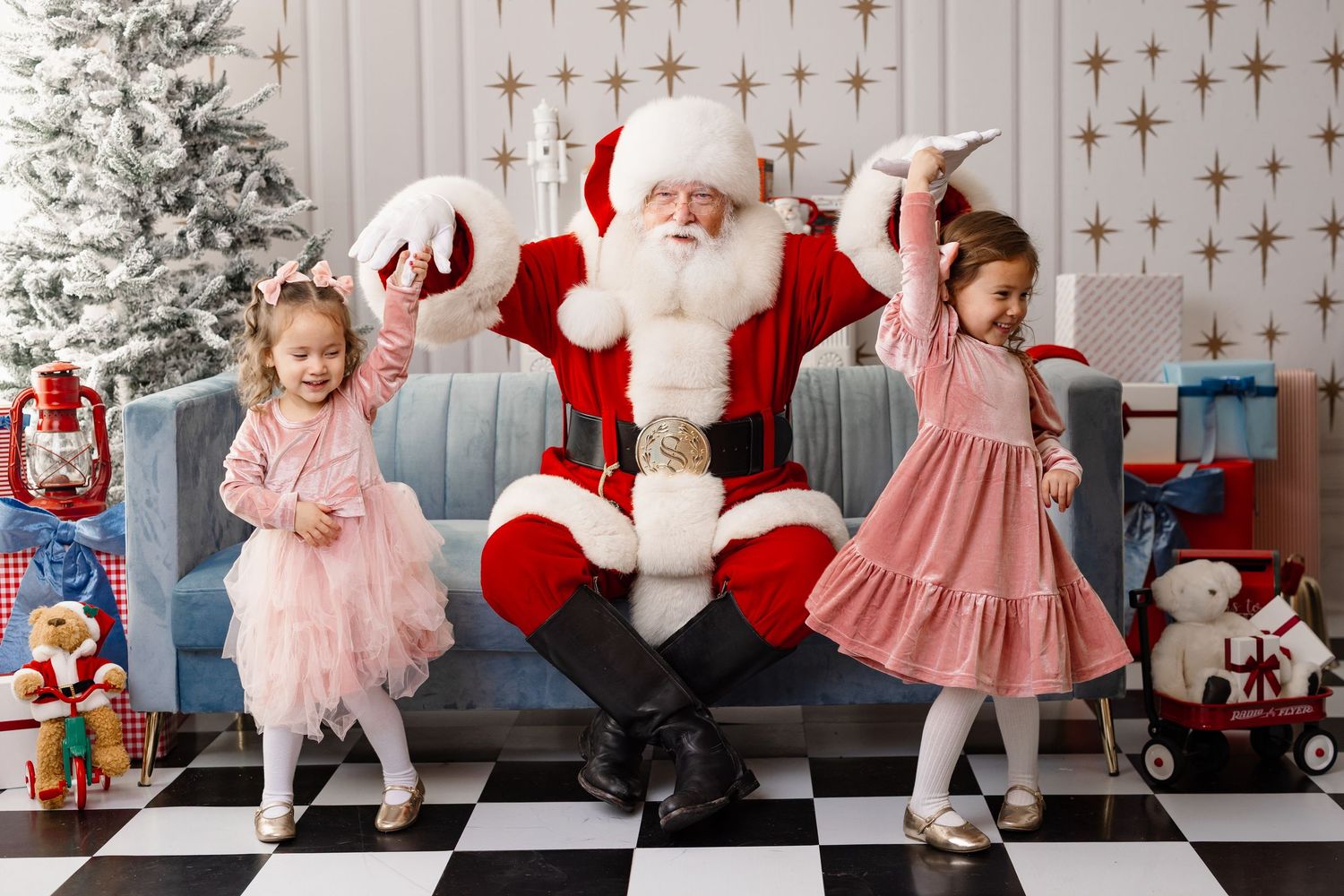 Two children in pink dresses dance joyfully beside Santa Claus on a black and white checkered floor with Christmas decorations.