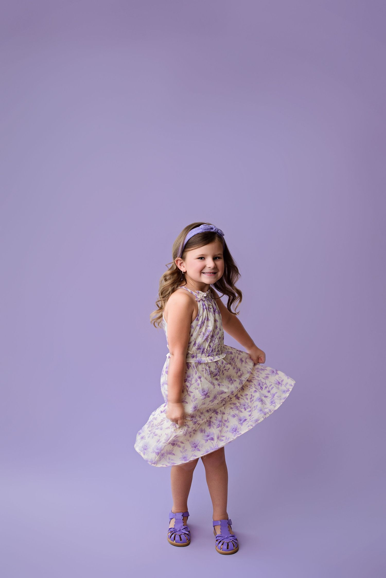 Small child wearing a floral dress twirls playfully against a purple backdrop while wearing purple sandals.