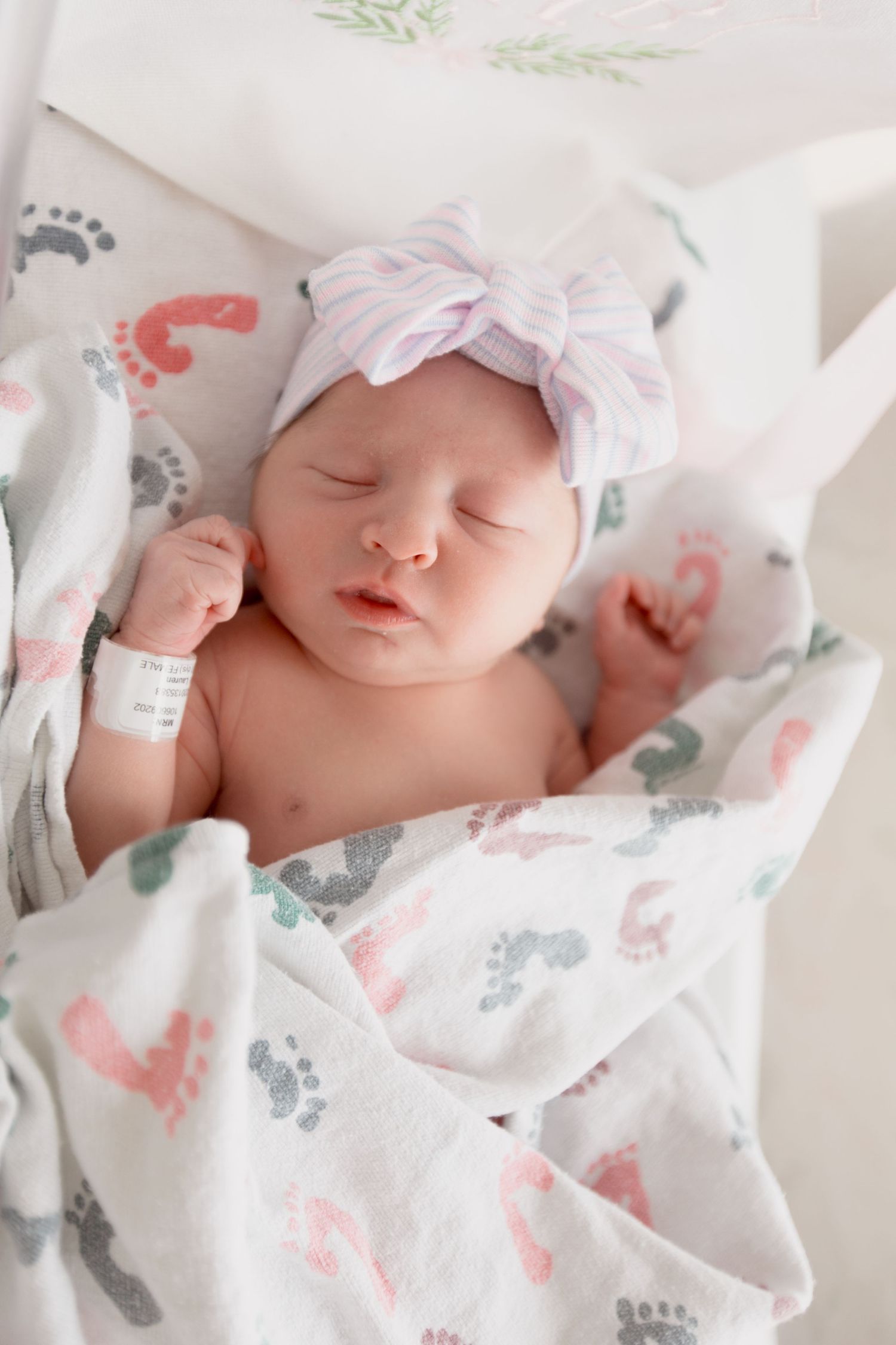 Peaceful newborn resting on dinosaur print swaddle blanket wearing pink bow headband during hospital photoshoot.