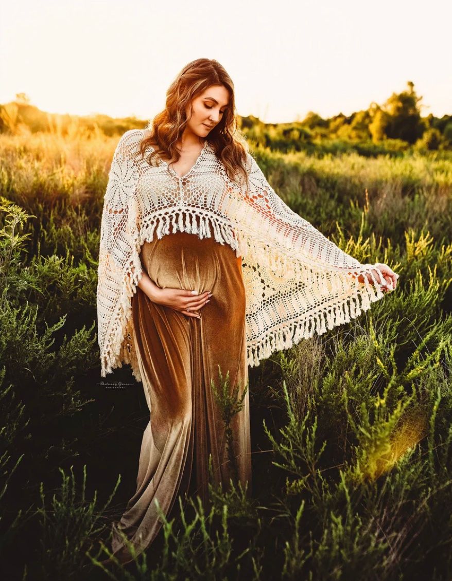 A woman in a brown velvet dress and cream lace shawl poses gracefully in a sunlit field at golden hour.