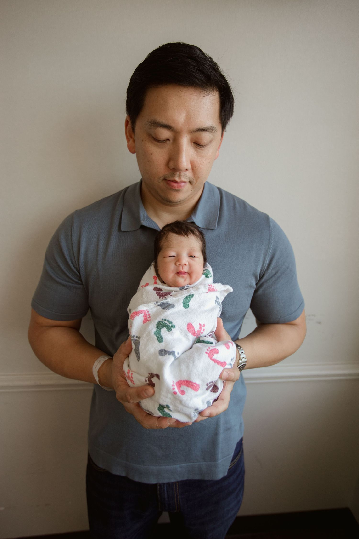 Dad in grey shirt holding newborn wrapped in patterned swaddle against neutral wall during Fresh 48 session in Beaumont hospital.