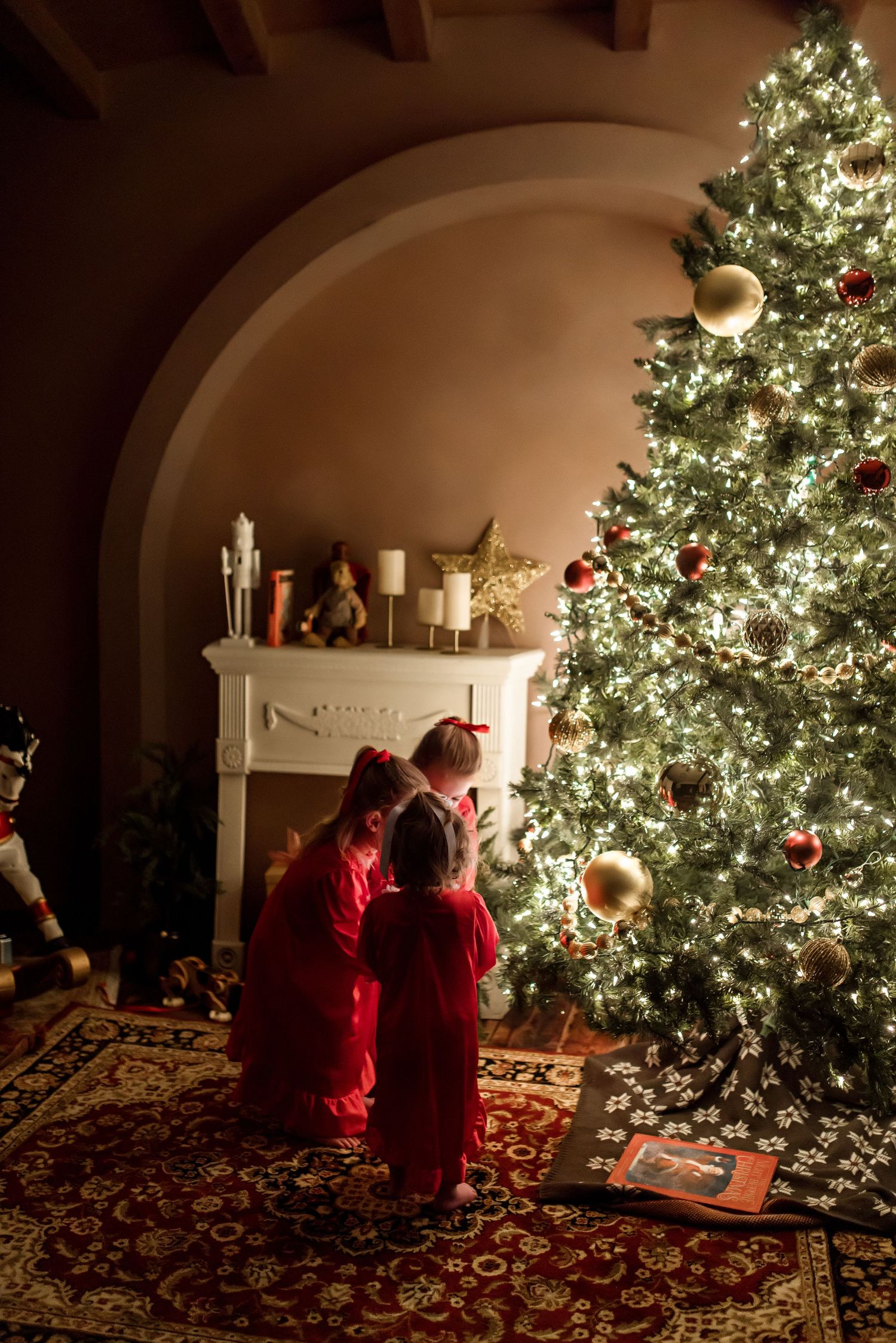 Two people in red admiring a beautifully lit Christmas tree beside a decorative fireplace.