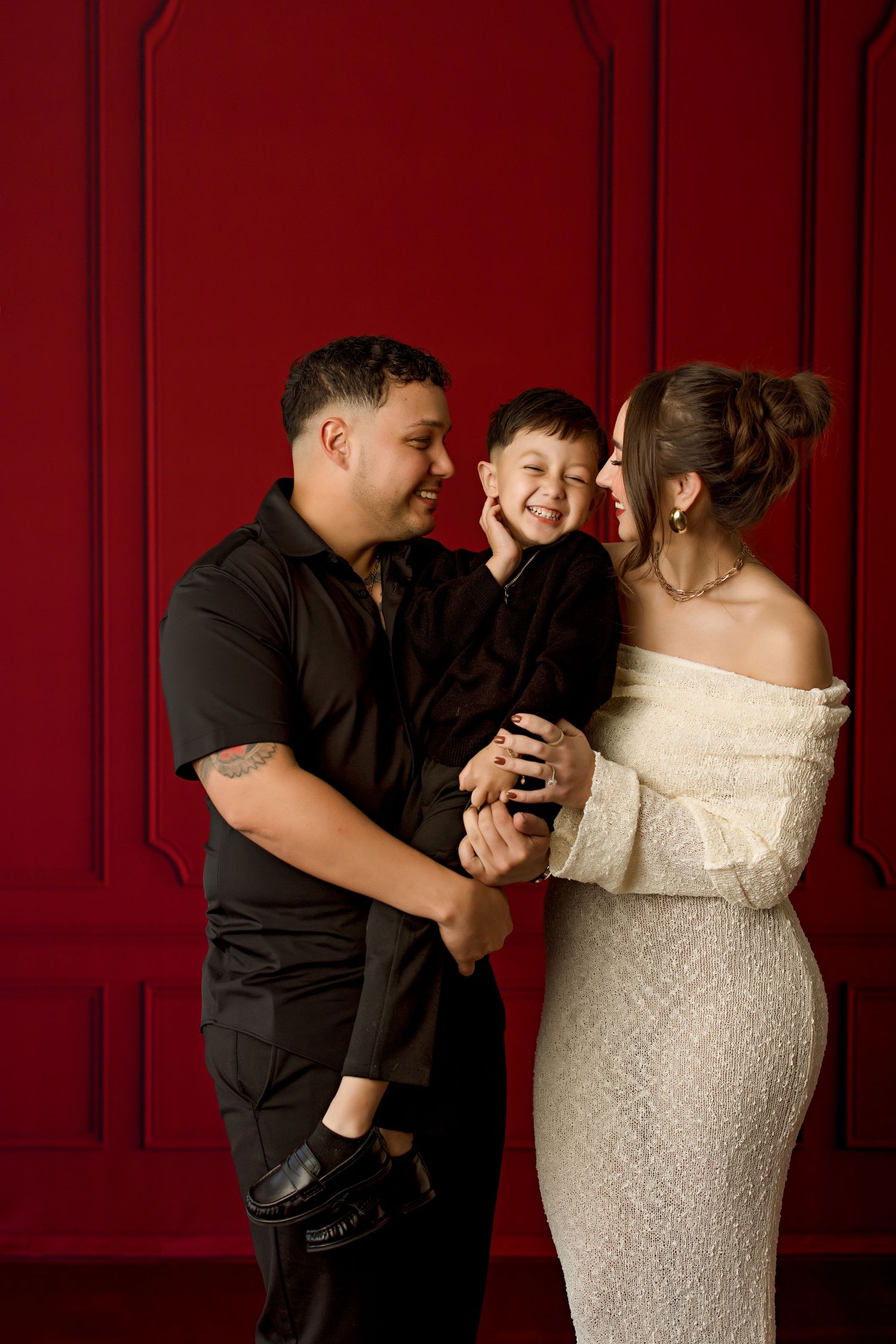 A couple in black and white attire share a joyful moment with their child against a deep red wall.