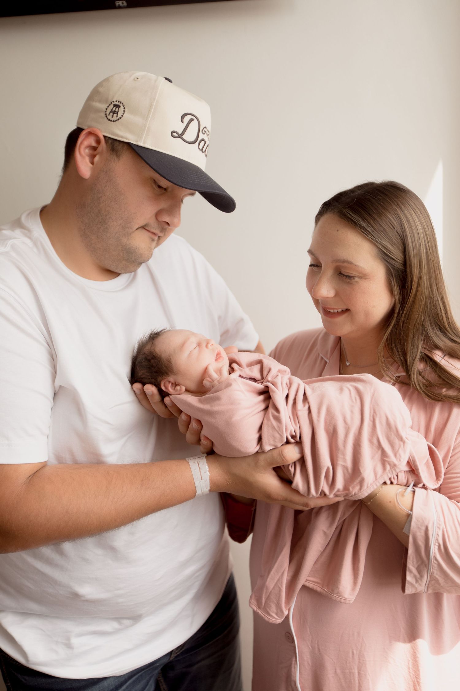 Parents in white and pink outfits lovingly hold their sleeping newborn baby wrapped in pink swaddle.