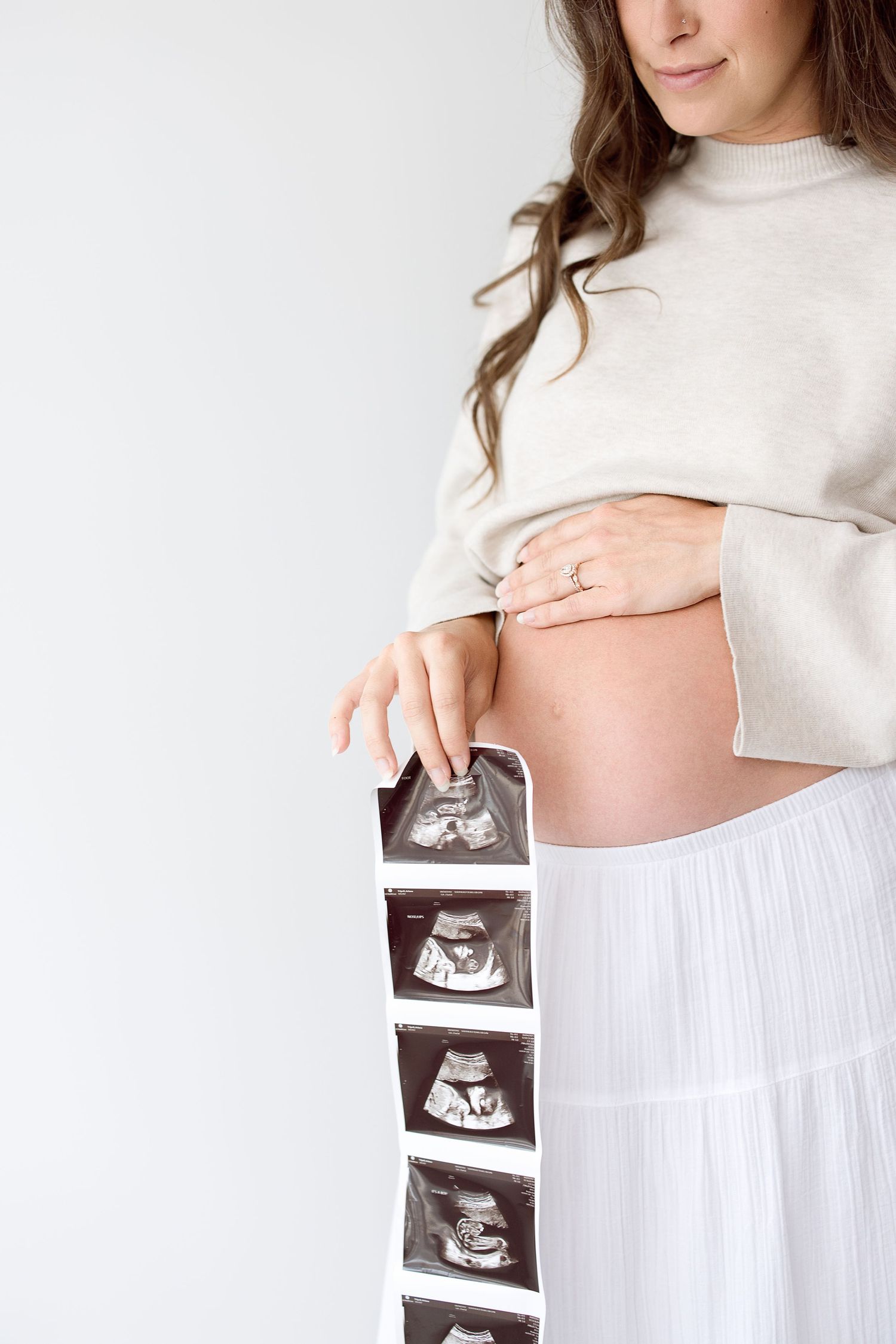 A pregnant woman in a cream turtleneck sweater holds ultrasound photos while posing against a white backdrop.