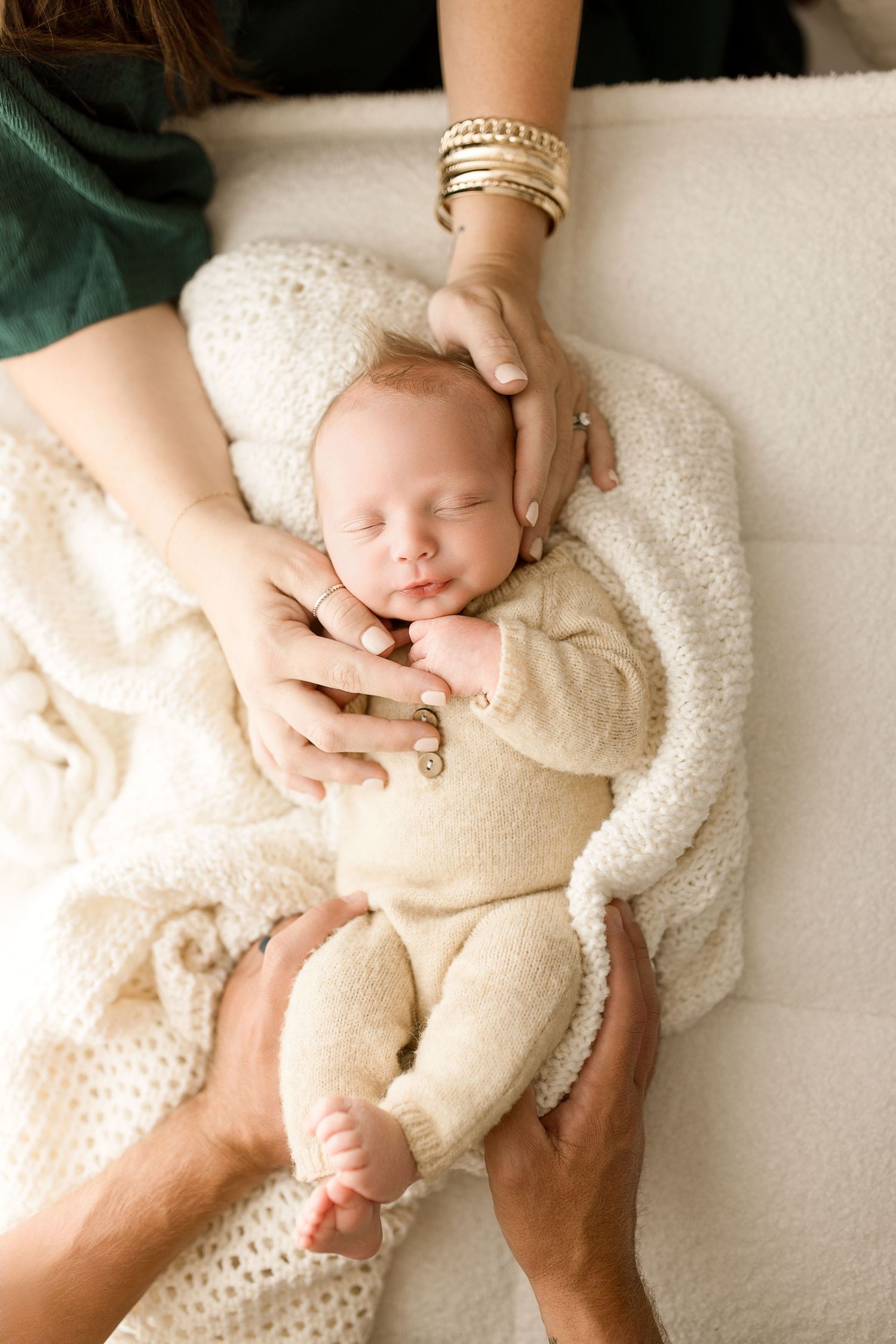 A series of gentle hands cradle a sleeping newborn baby wearing a cream-colored knit outfit on a white blanket.