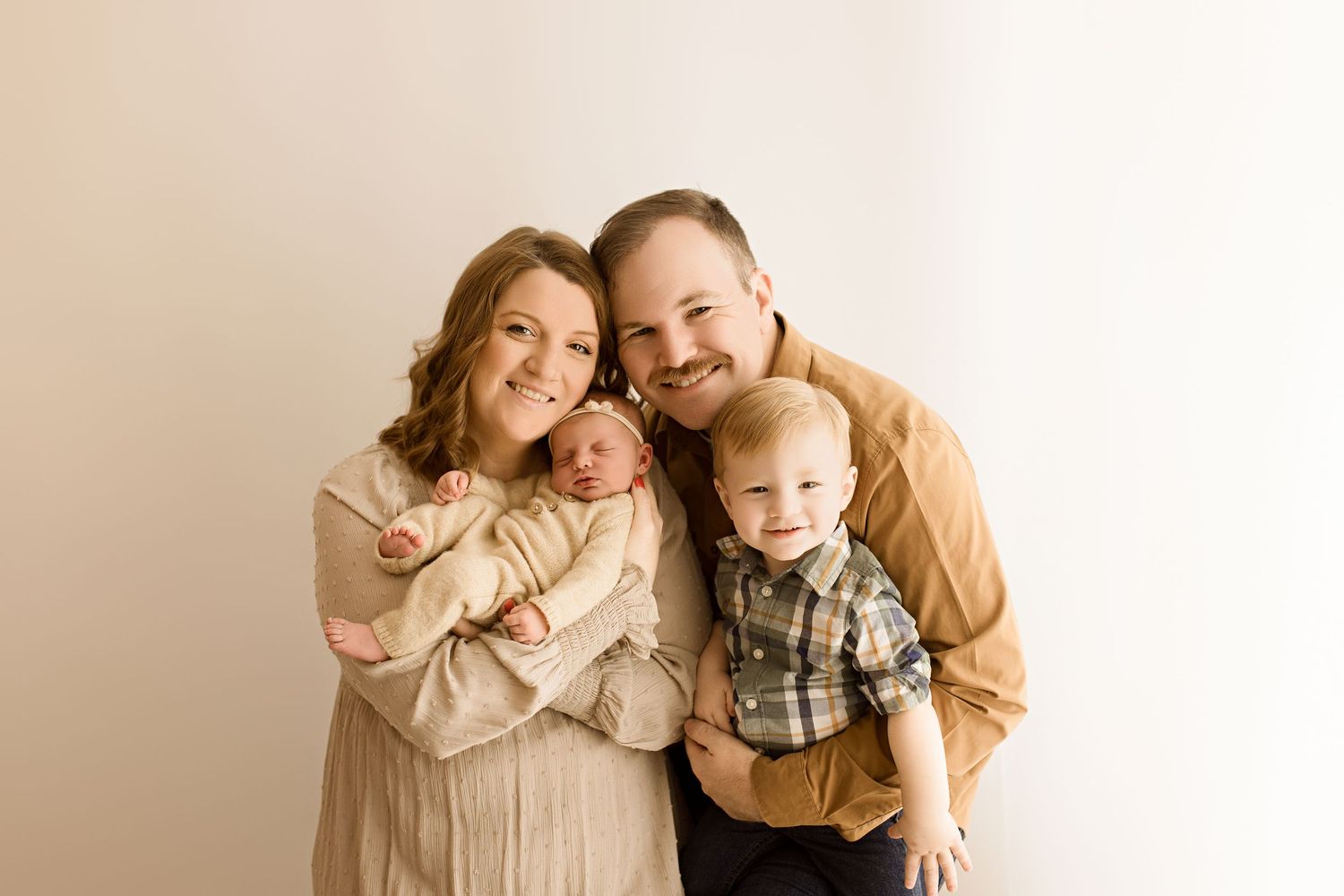 Family of three poses together against cream wall wearing coordinated neutral colored outfits.