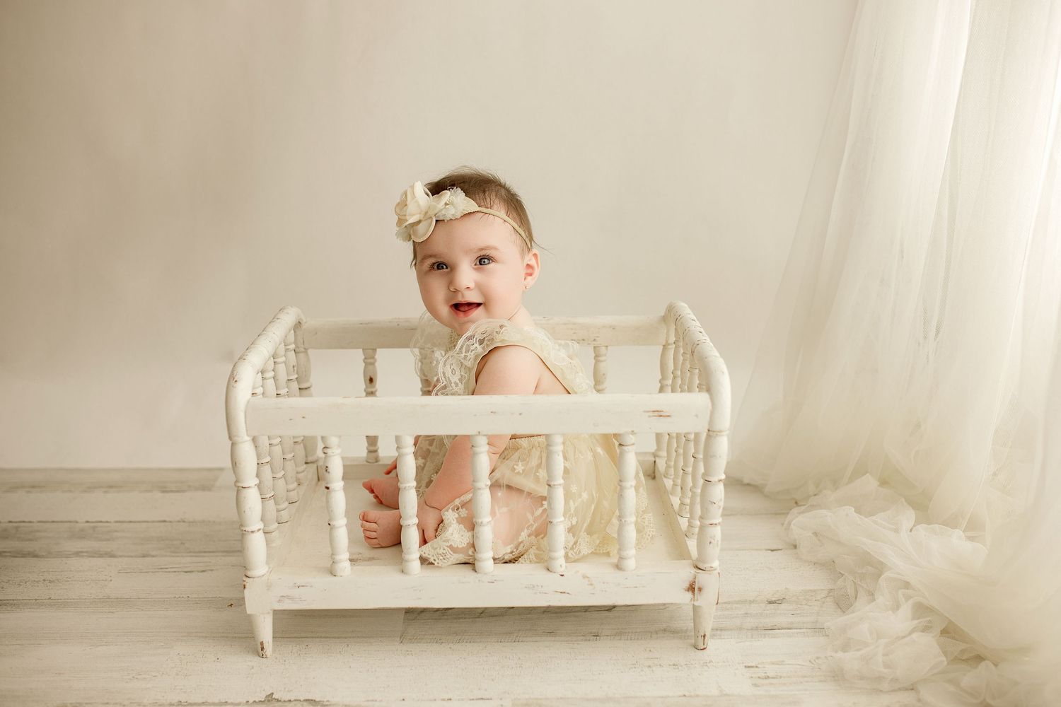 Small baby wearing a light pink lace romper outfit laughs while posing in a white vintage daybed during studio photo shoot.