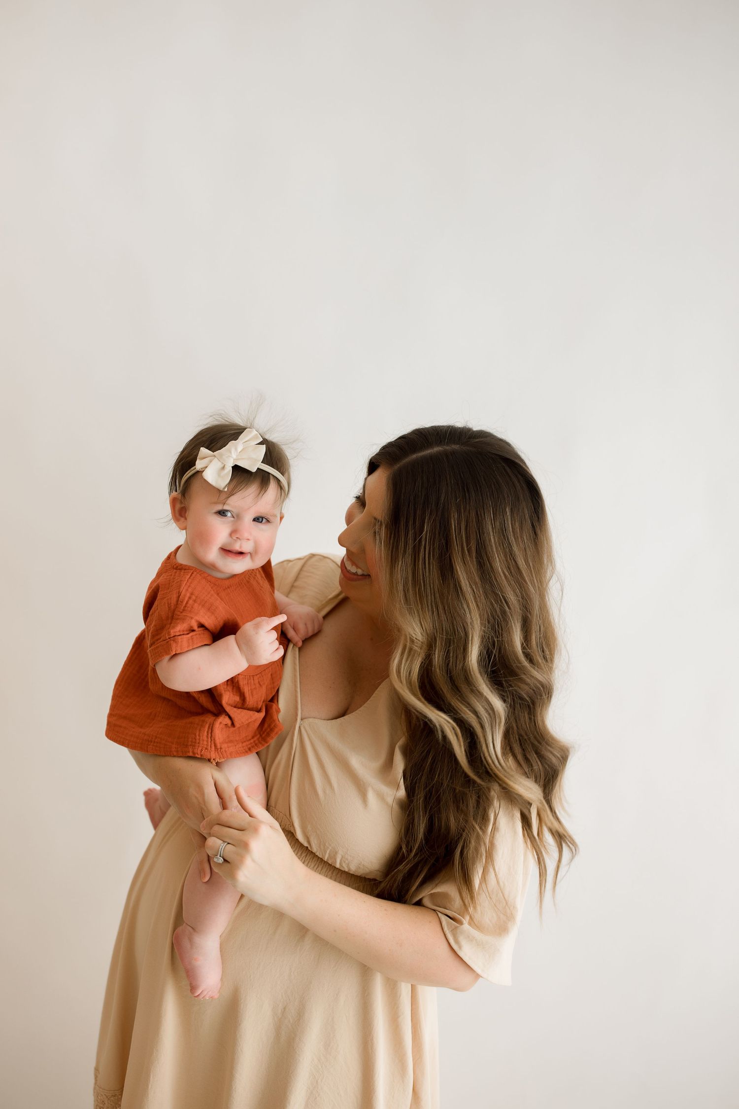 Parent and baby in matching beige outfits share tender moments in minimalist portrait series against white background.