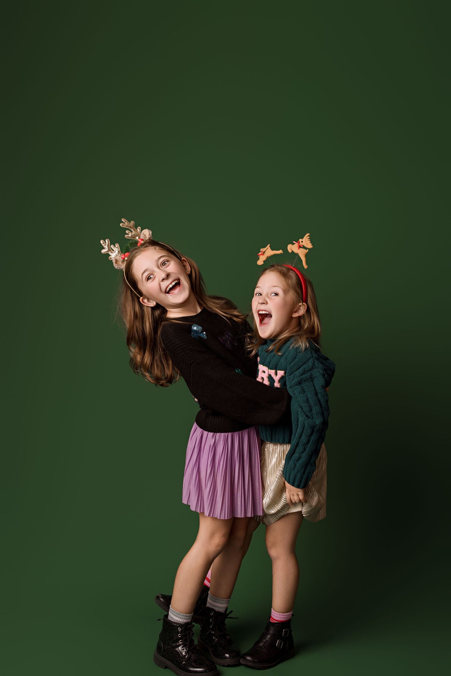 Two children wearing festive holiday attire laugh and pose playfully against a dark green background.