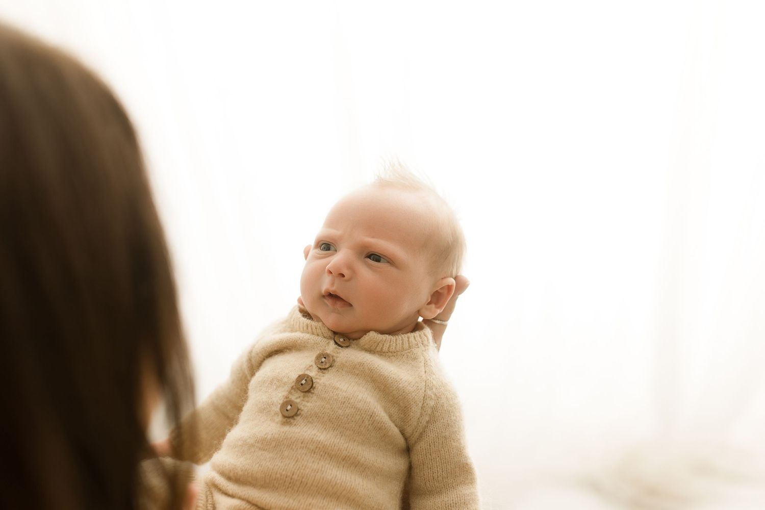 Baby wearing a beige knit sweater sits looking up against a bright white background.