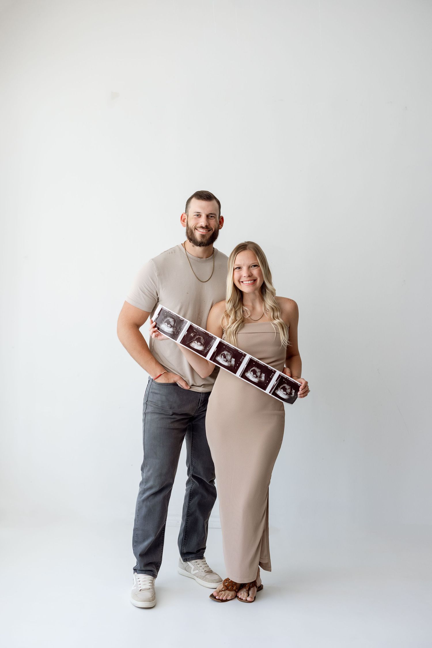 A couple in matching beige outfits poses together during a pregnancy announcement photoshoot with ultrasound photos.