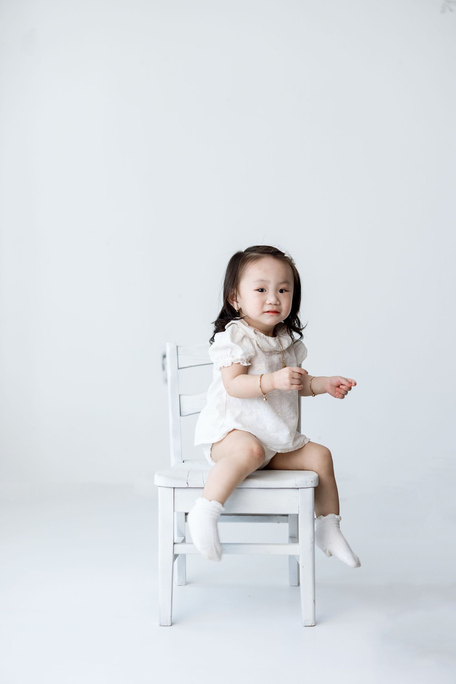 Baby in white sitting on a small white chair while looking and pointing away from the camera against white background.