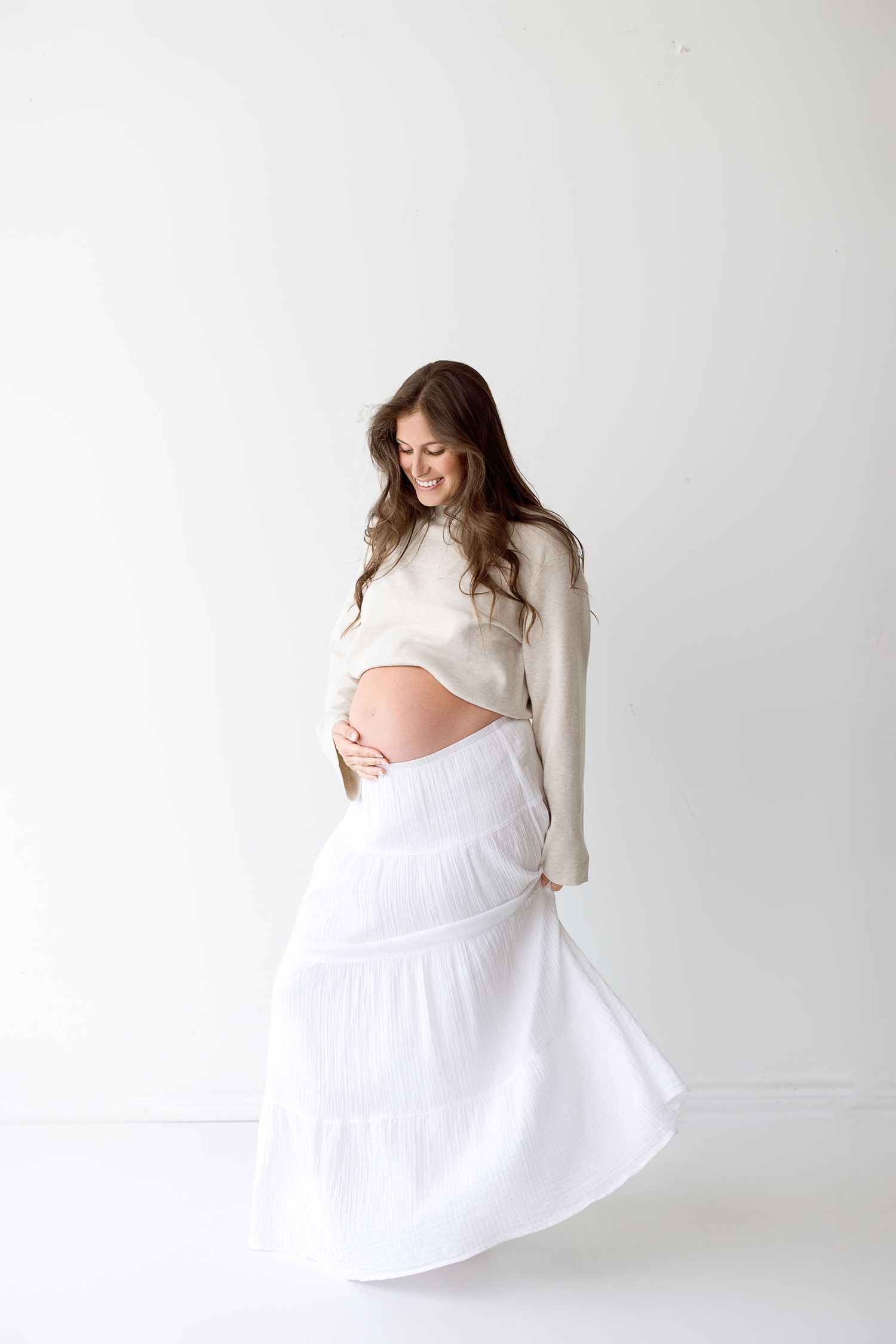 Maternity photoshoot featuring flowing white skirt and cream sweater against minimalist white backdrop.