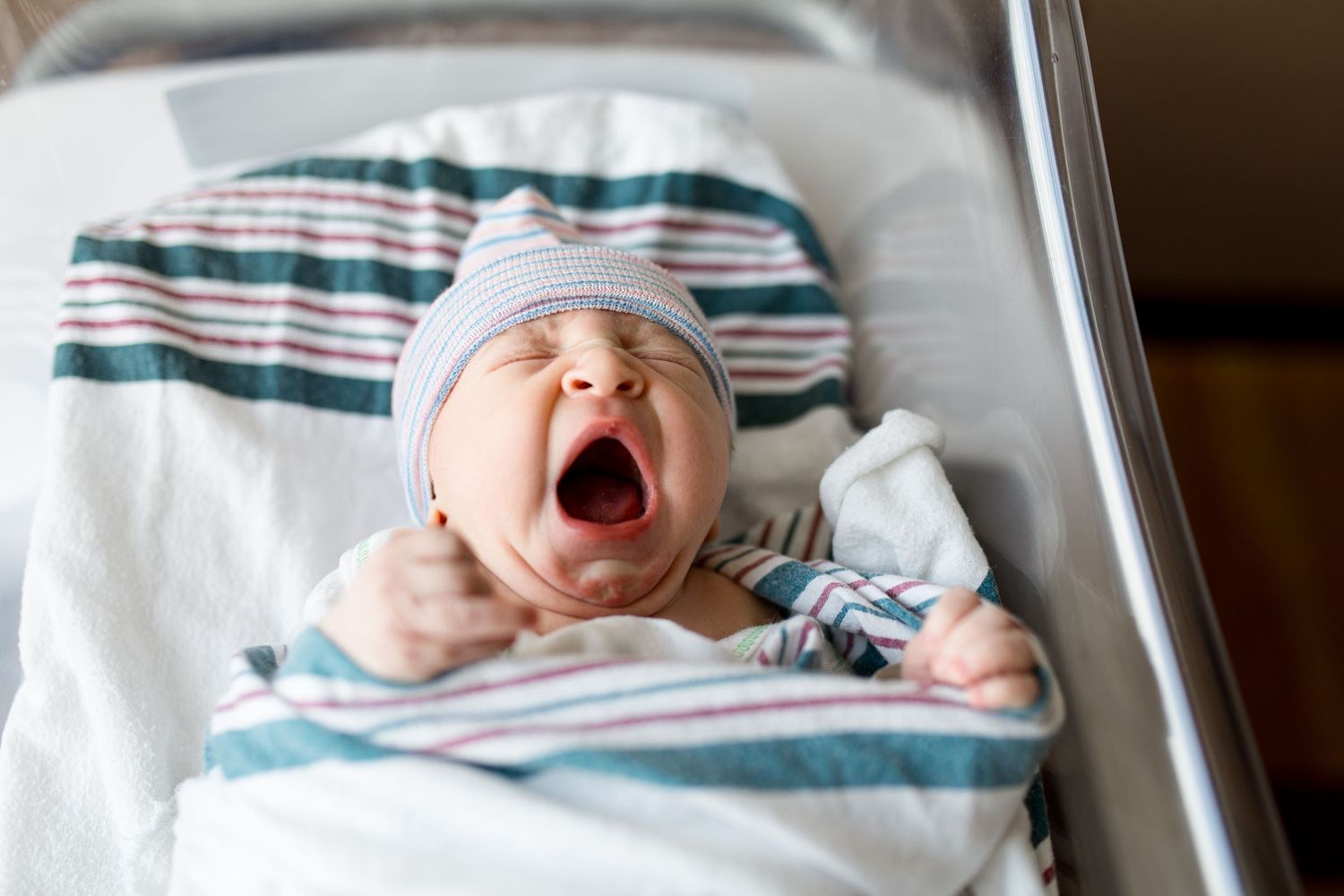 Newborn baby yawning in hospital crib, wrapped in striped blanket during Fresh 48 session with Bree Elle Photography at Baptist Hospital.