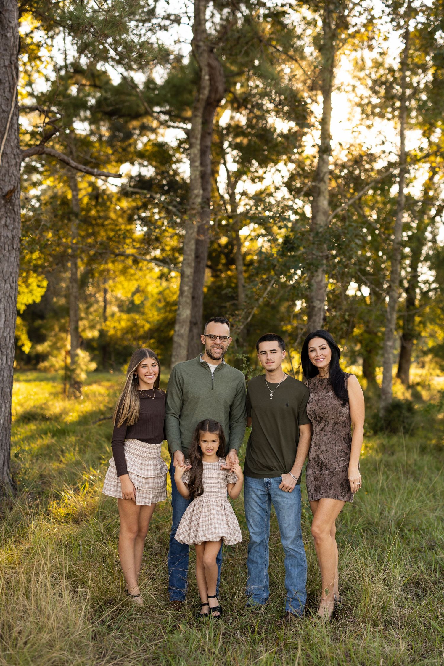 A group of people stand together in a grassy field at sunset with tall pine trees in the background.